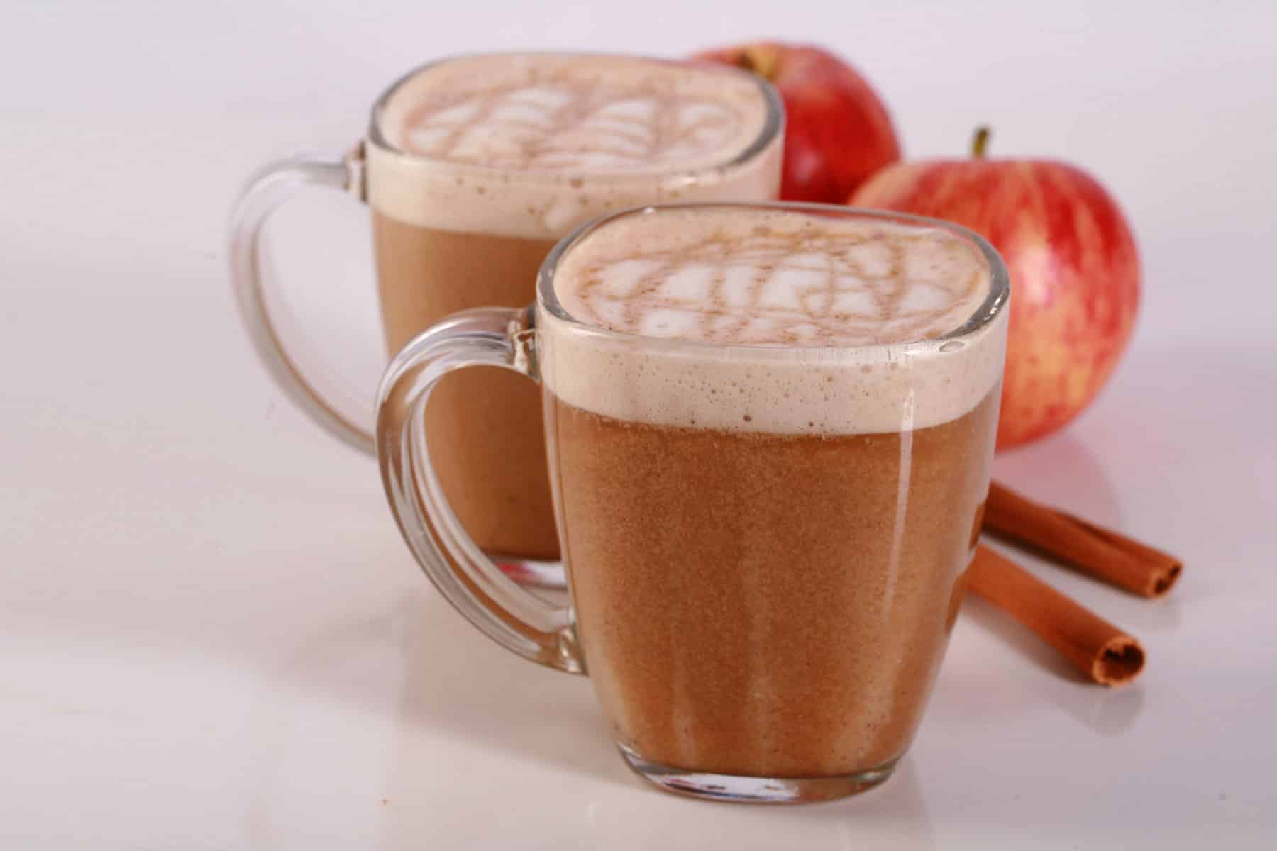 2 glass mugs of apple crisp macchiatos, with cinnamon sticks and apples next to them.