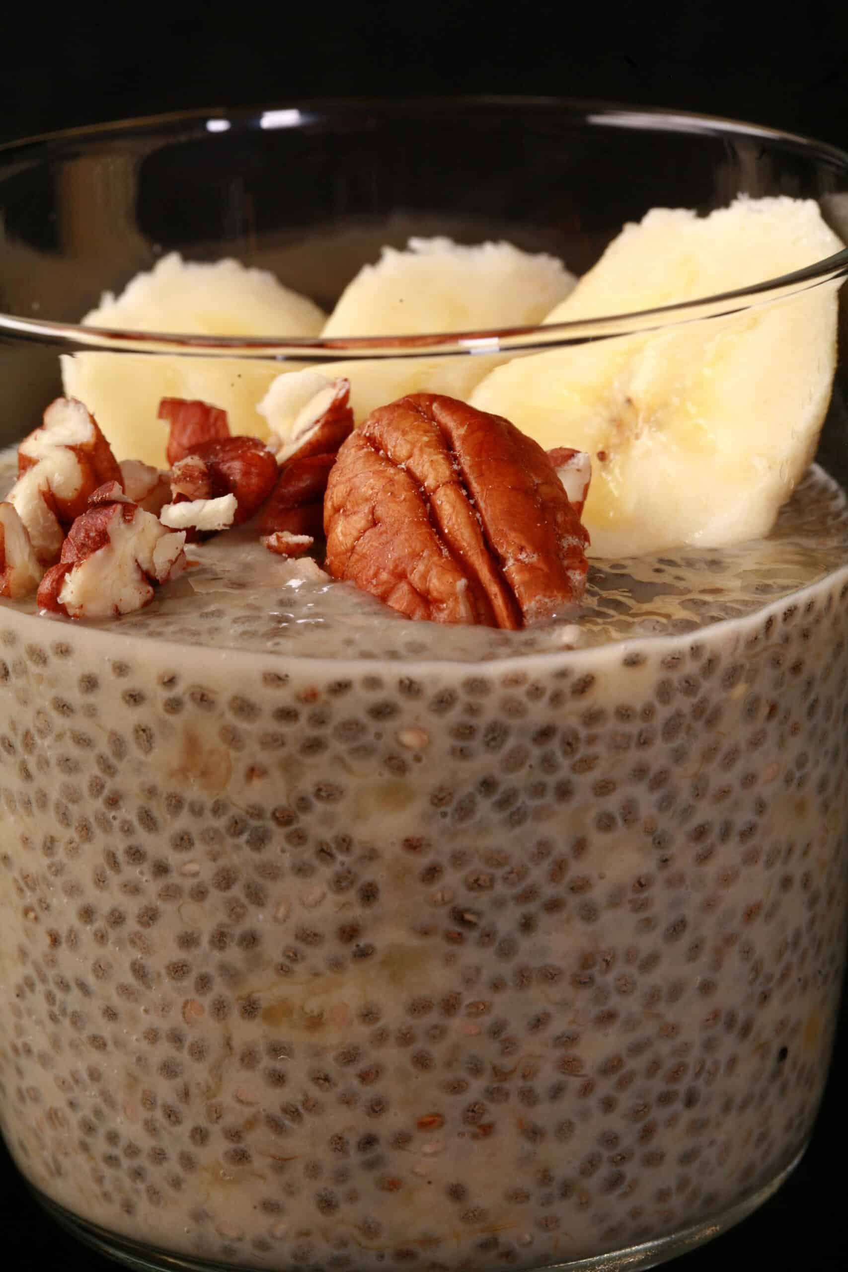 A close up view of a glass of banana chia pudding, topped with banana slices and pecans.