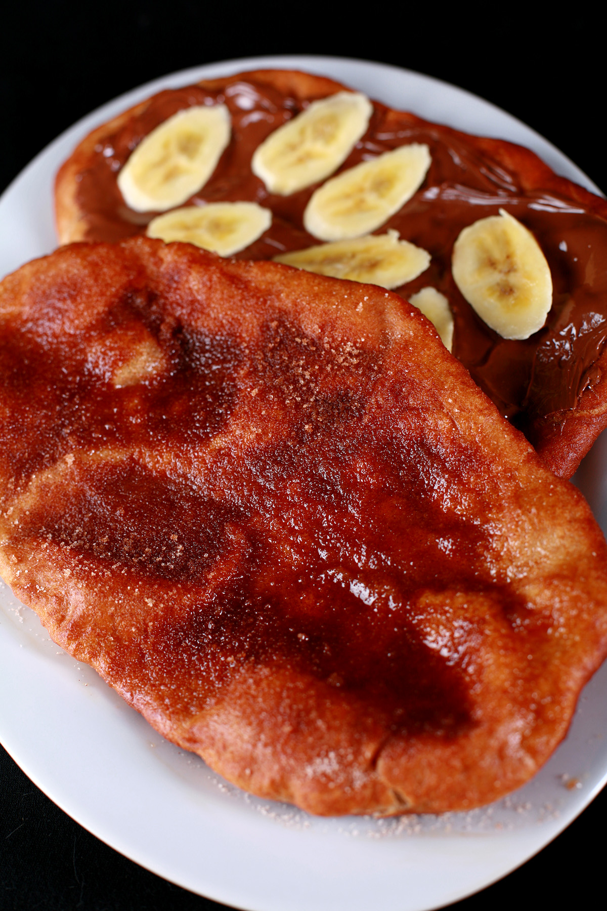 Two gluten-free fried pastries - made from this Gluten-free Beaver Tail recipe - are arranged on a plate. Both are oblong pieces of fried dough. One is topped with cinnamon sugar, the other is spread with Nutella and topped with sliced banana.