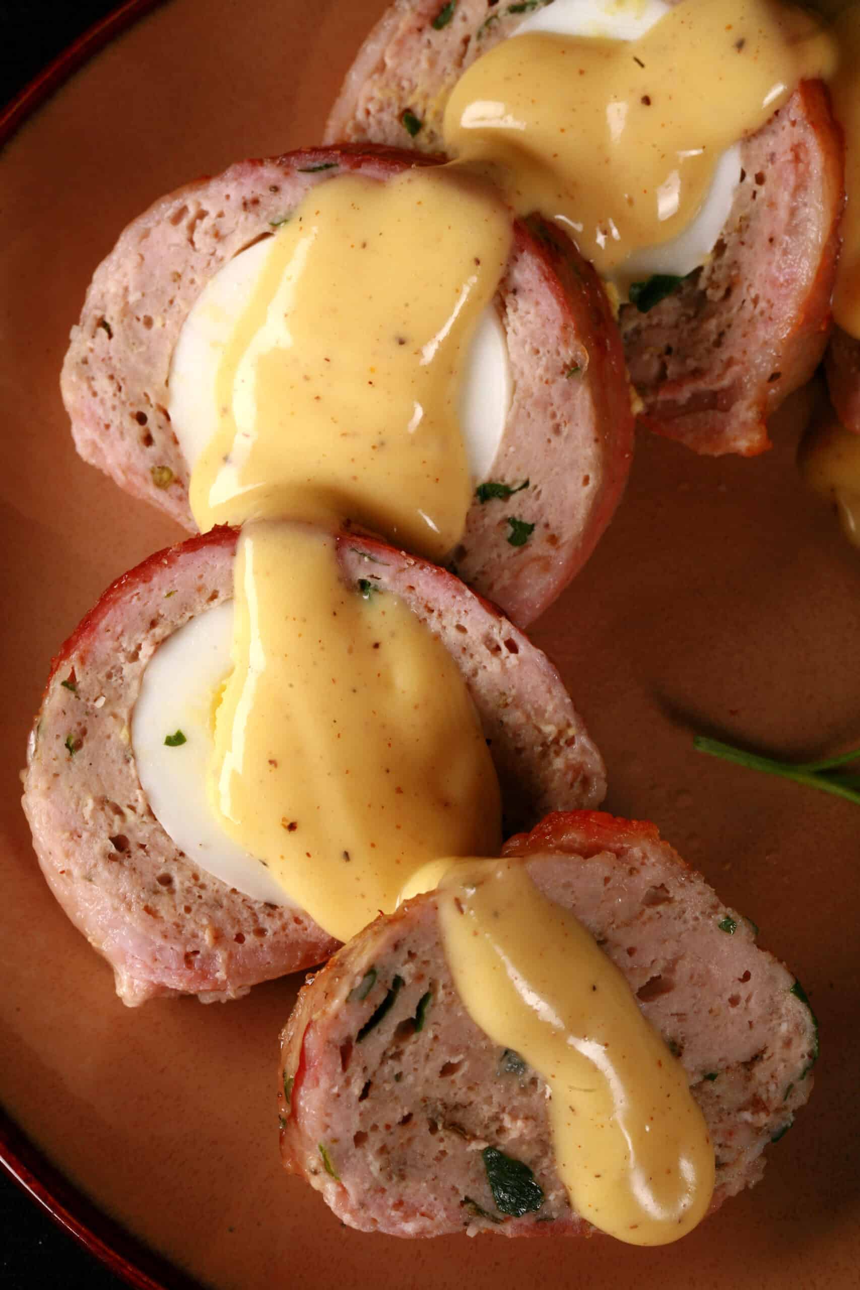 A sliced individual breakfast meatloaf, wrapped in bacon, stuffed with a hard boiled egg, and topped with hollandaise sauce.