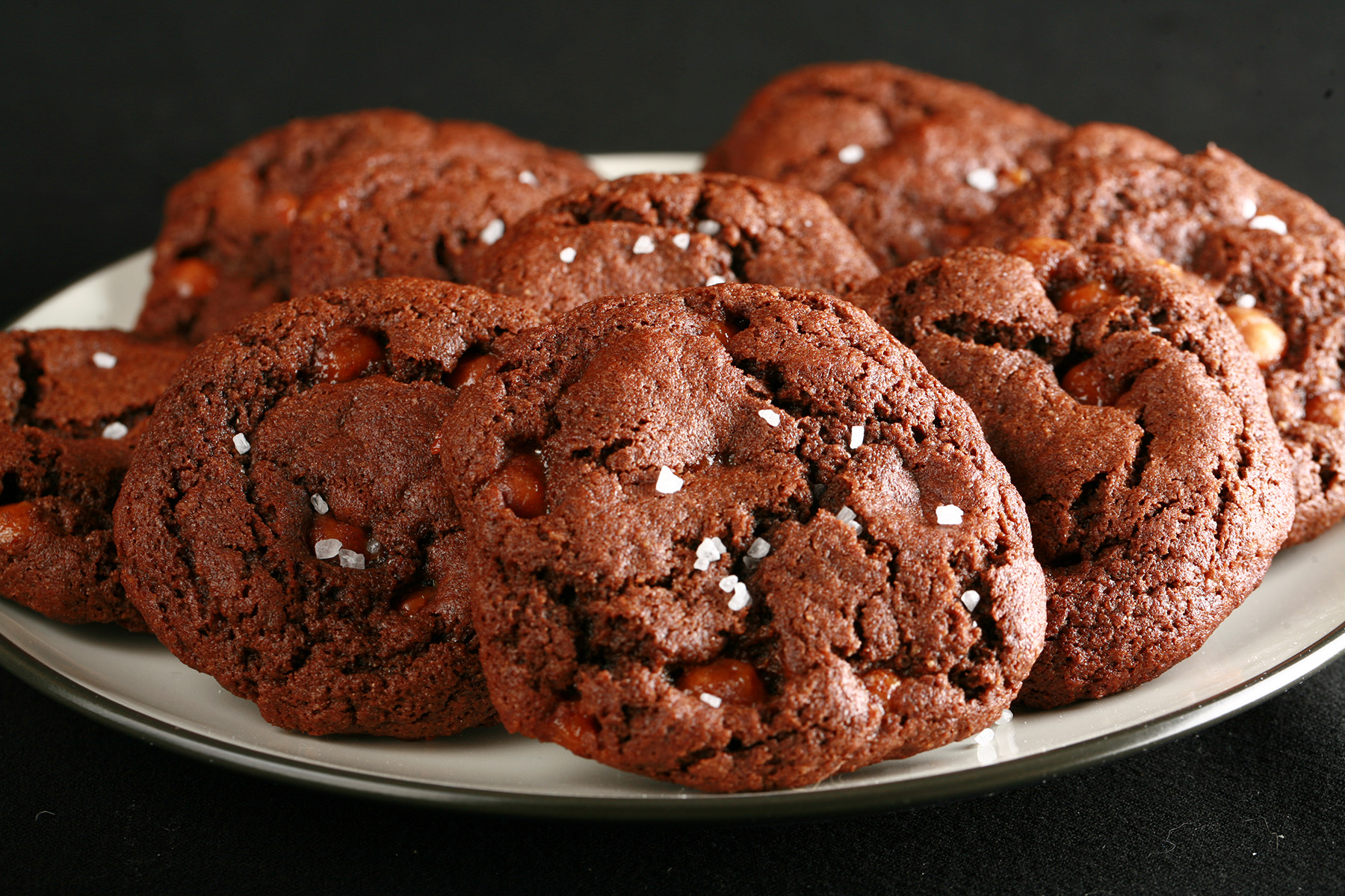 A small white plate, piled with Chewy, Gluten-Free Chocolate Cookies. Bits of caramel are visible within the cookies, and each is topped with a sprinkle of sea salt.
