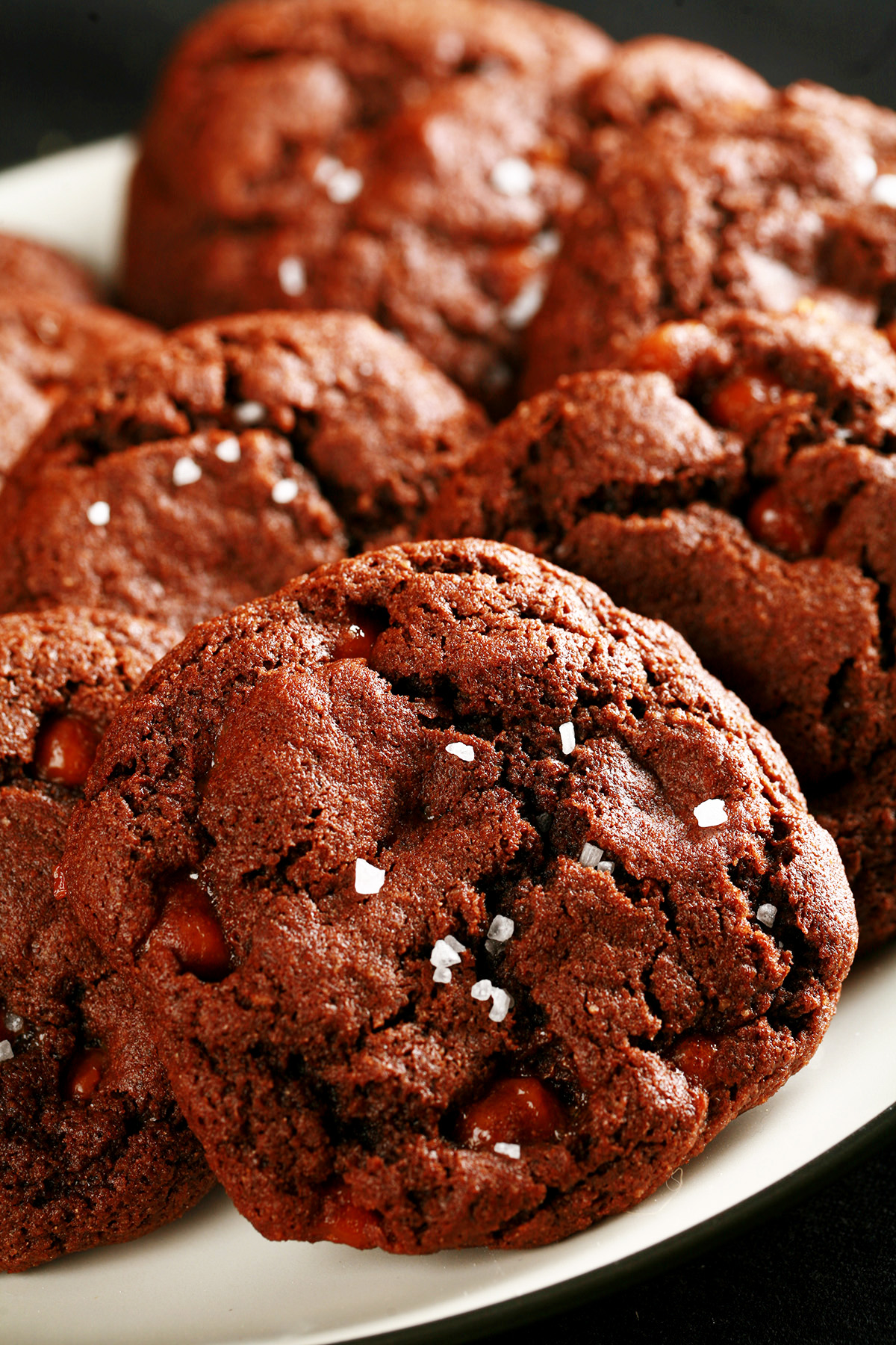 A small white plate, piled with Chewy, Gluten-Free Chocolate Cookies. Bits of caramel are visible within the cookies, and each is topped with a sprinkle of sea salt.