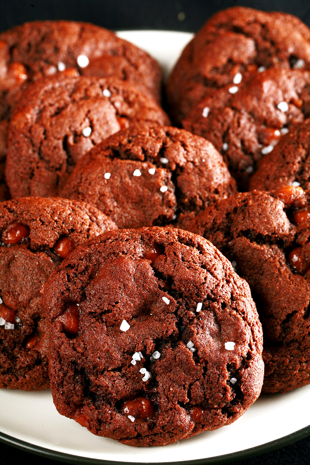 A small white plate, piled with Chewy, Gluten-Free Chocolate Cookies. Bits of caramel are visible within the cookies, and each is topped with a sprinkle of sea salt.