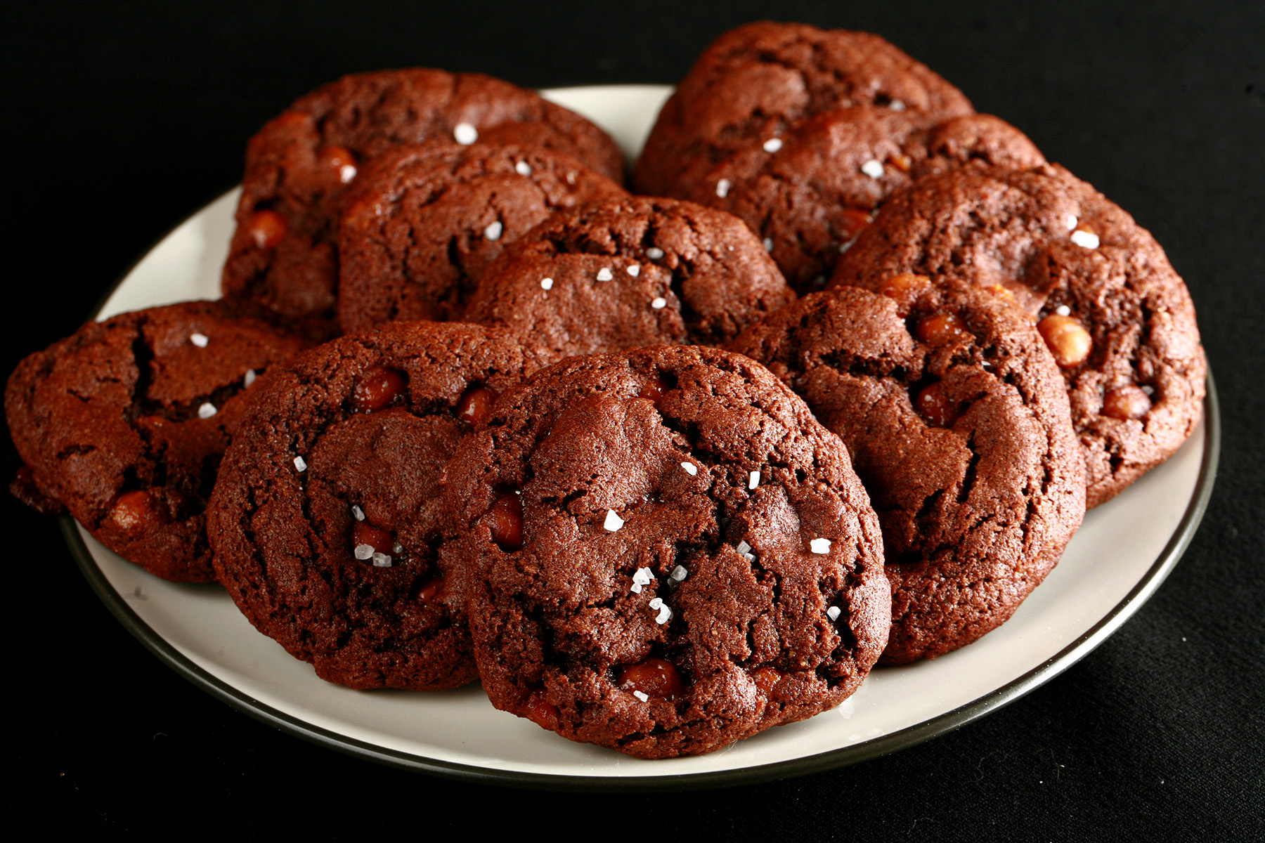 A small white plate, piled with Chewy, Gluten-Free Chocolate Cookies. Bits of caramel are visible within the cookies, and each is topped with a sprinkle of sea salt.