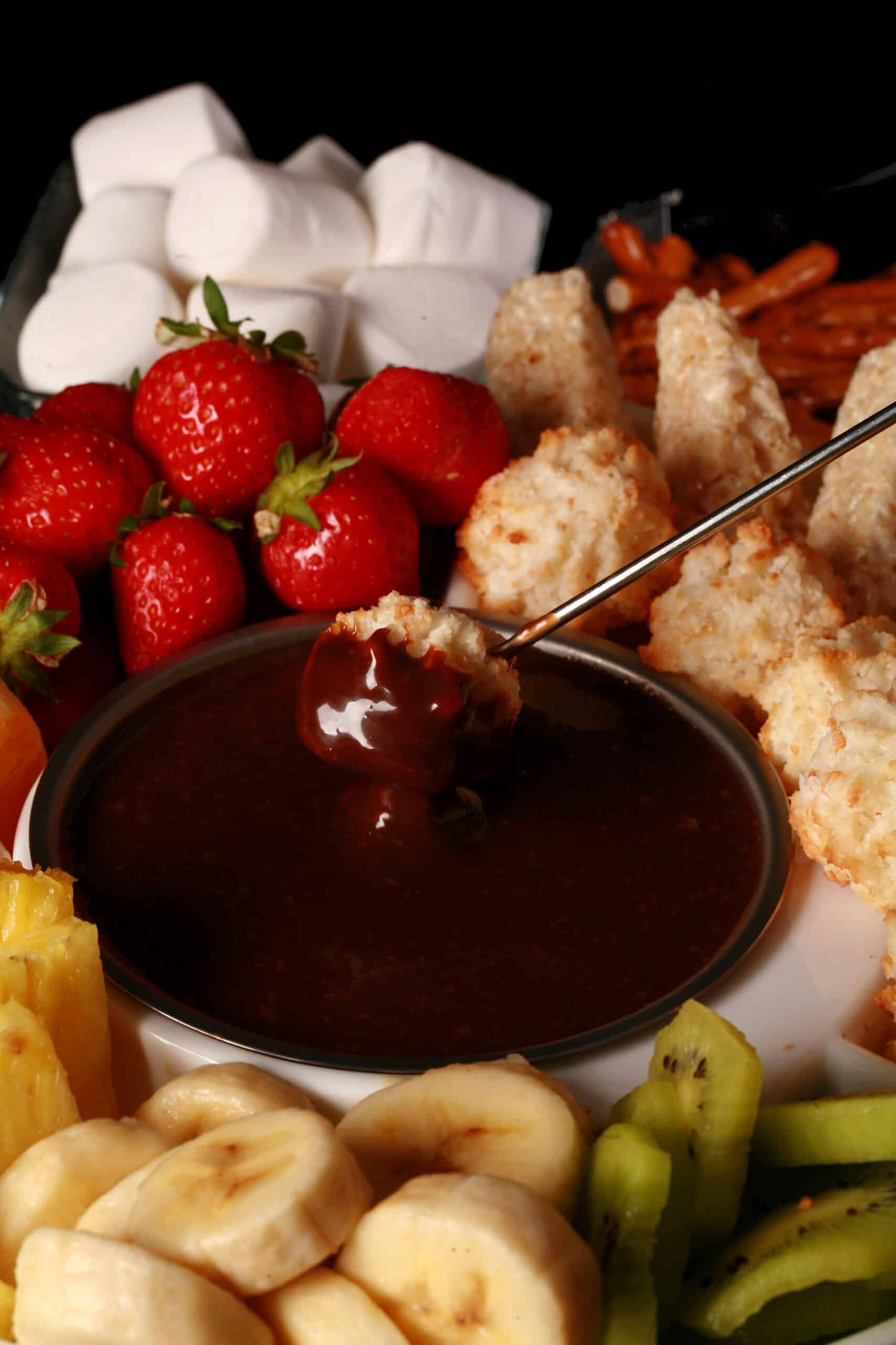 A coconut macaroon being dipped into chocolate fondue, surrounded by pineapple chunks, banana slices, kiwi slices, coconut macaroons, oranges, pretzels, marshmallows, and more.