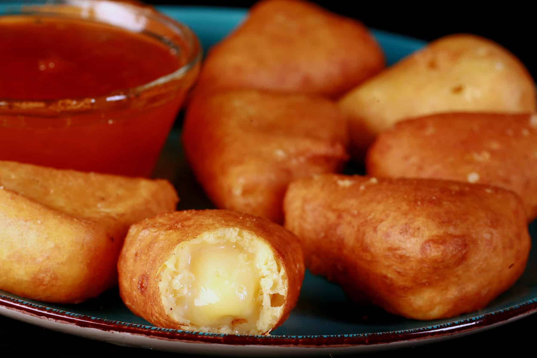 A plate of battered gluten free deep fried brie cheese wedges, around a bowl of apricot preserves. One has a bite, with brie oozing out.