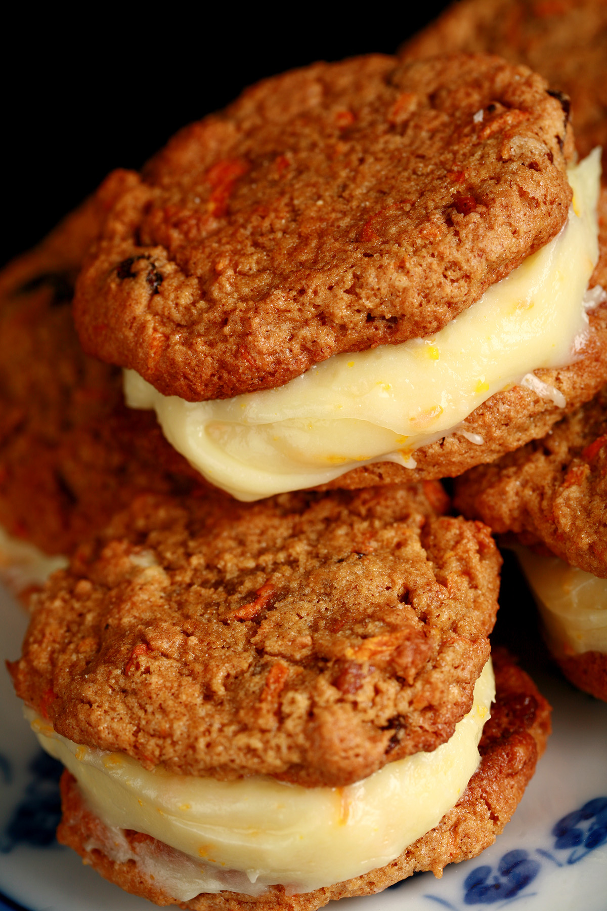 "A small blue and white plate with a stack of gluten-free carrot cake cookies on it. The filling is ivory coloured and has flecks of orange zest throughout.