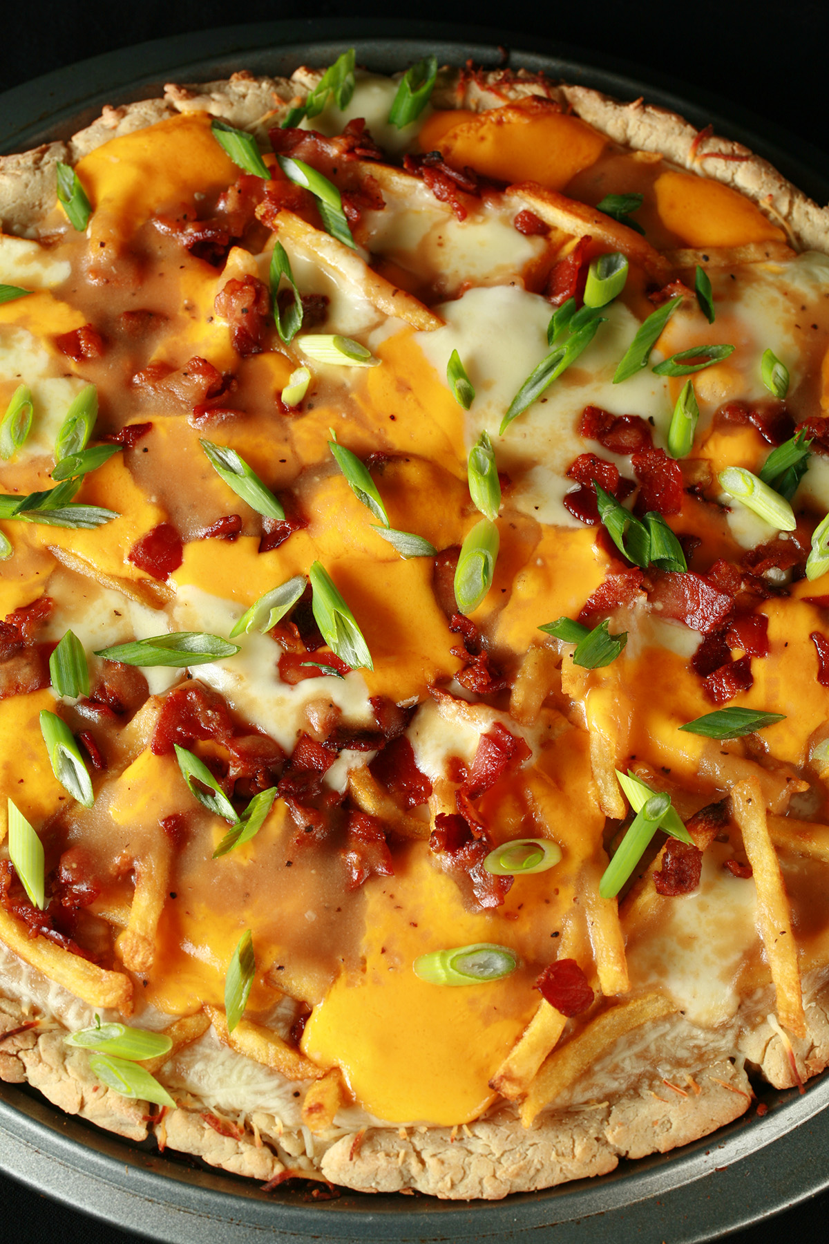A close up view of a gluten-free bacon poutine pizza - A pizza crust topped with gravy, fries, cheese curds, chredded cheese, bacon, and green onions!