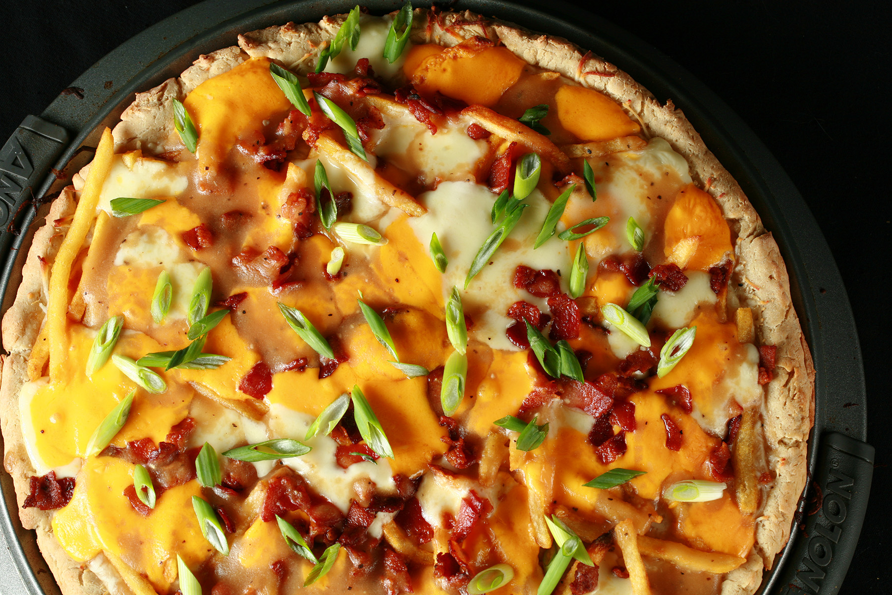 A close up view of a gluten-free poutine pizza - A pizza crust topped with gravy, fries, cheese curds, chredded cheese, bacon, and green onions!