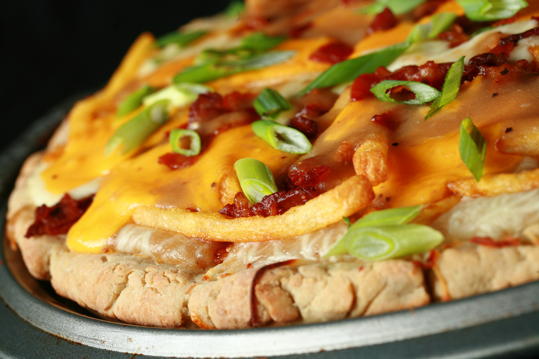 A close up view of a gluten-free poutine pizza - A pizza crust topped with gravy, fries, cheese curds, chredded cheese, bacon, and green onions!
