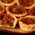 A brown plate is crowded with gluten-free buttertarts - mini tarts with a gooey sugar filling and raisins.