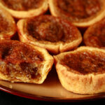 A brown plate is crowded with gluten-free buttertarts - mini tarts with a gooey sugar filling and raisins.