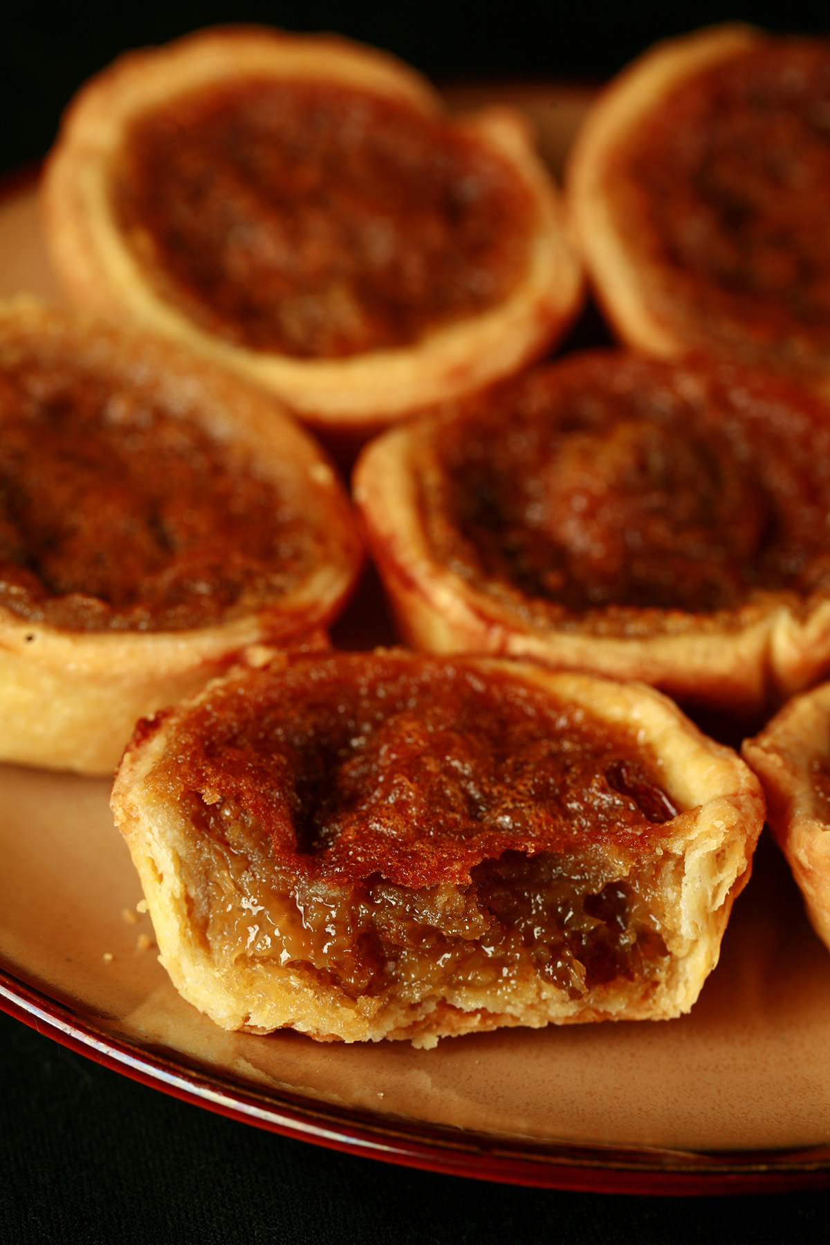 A brown plate is crowded with gluten-free buttertarts - mini tarts with a gooey sugar filling and raisins