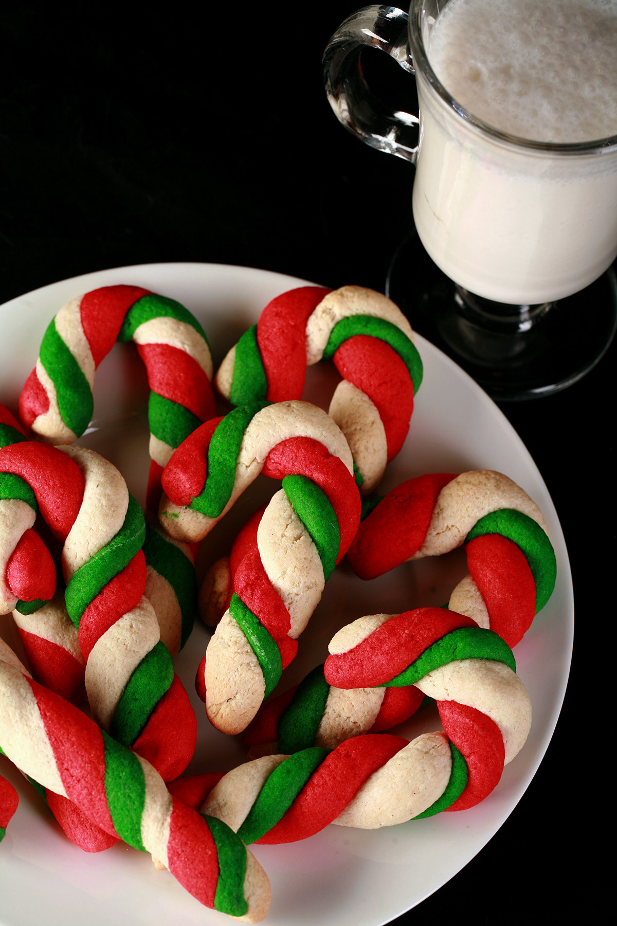 "Gluten-Free Candy Cane cookies - candy cane shaped cookies, made from lengths of red, green, and white twisted dough - on a plate. There is a glass of milk behind the plate.
