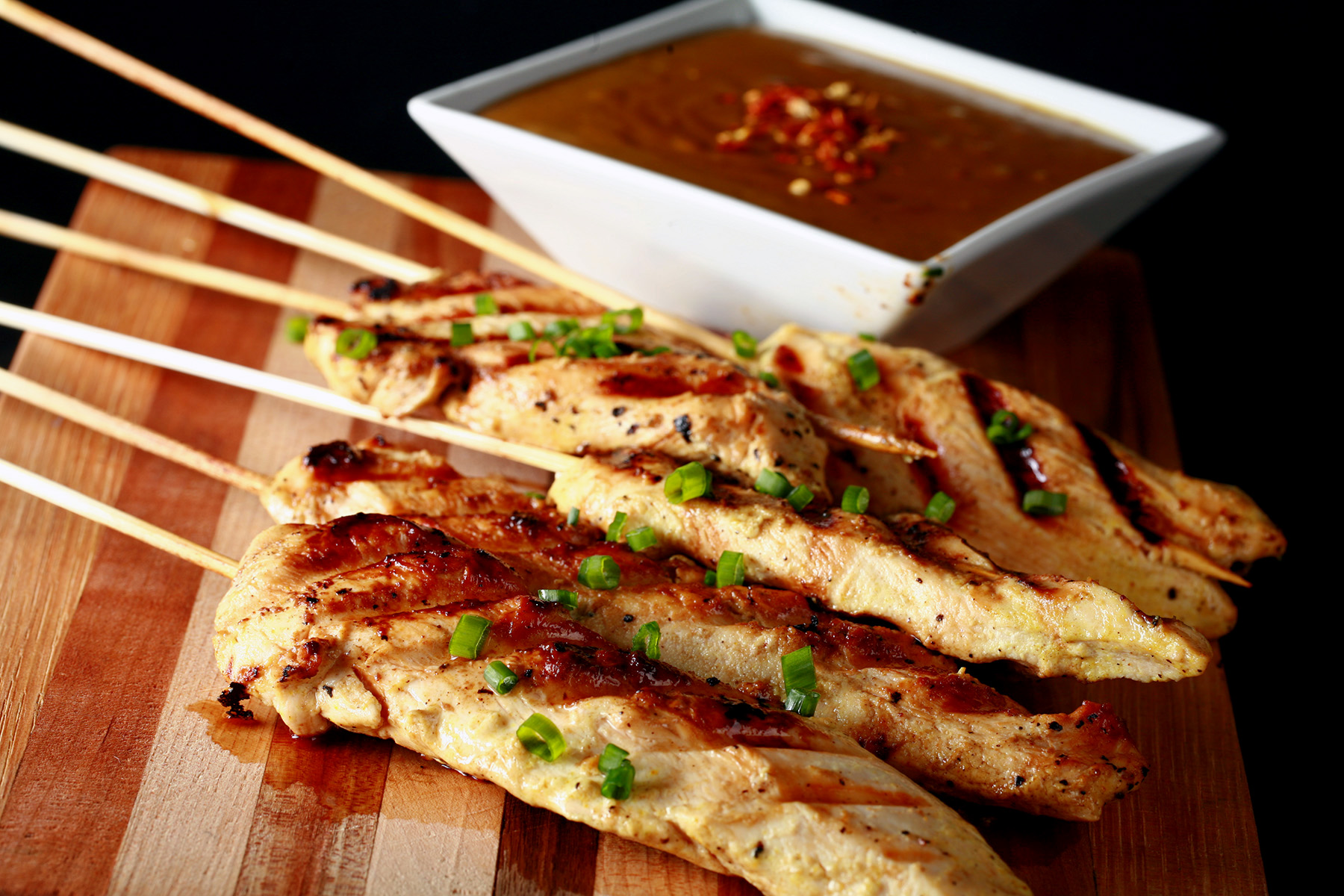 Several skewers of gluten-free chicken satay on a striped cutting board. They are sprinkled with green onions and sitting next to a bowl of peanut sauce.