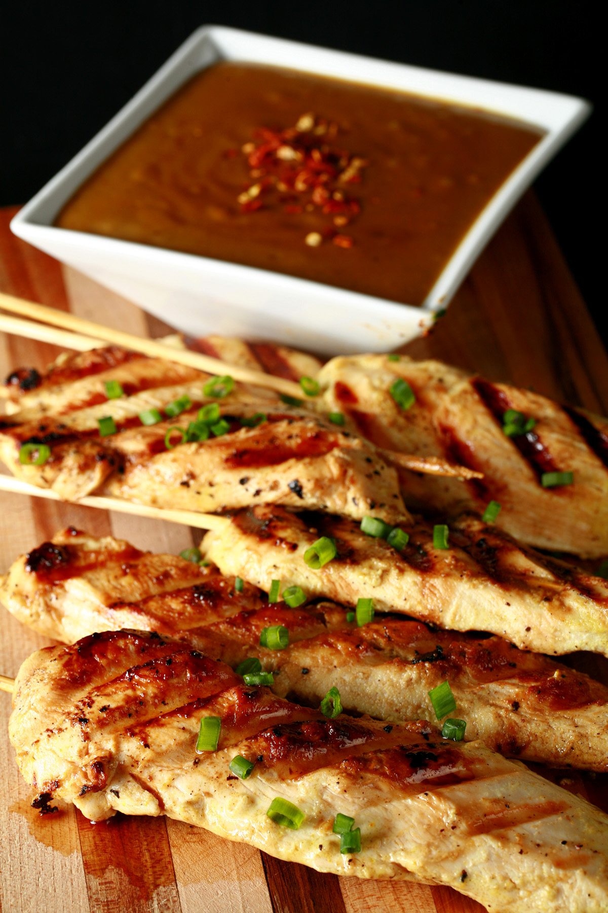 Several skewers of gluten-free chicken satay on a striped cutting board. They are sprinkled with green onions and sitting next to a bowl of peanut sauce.
