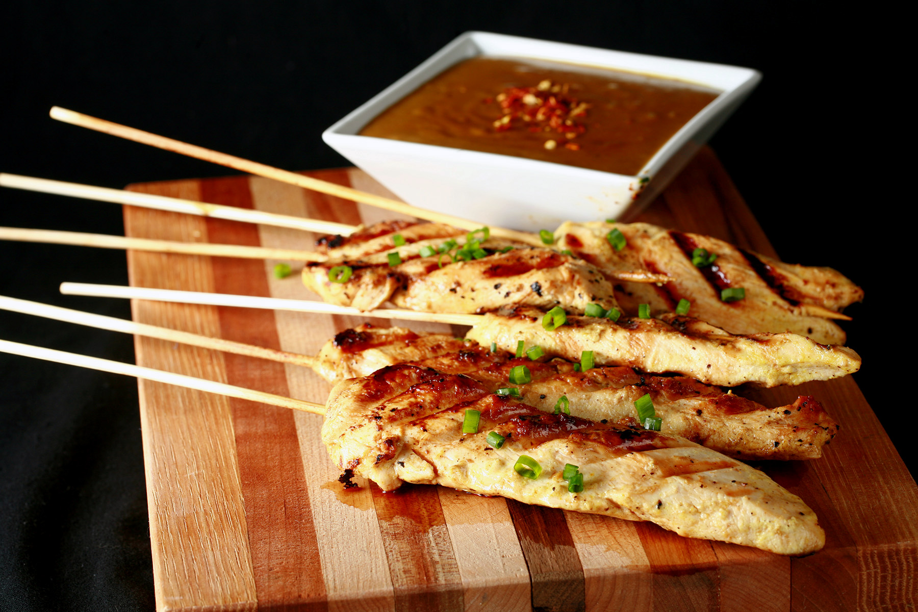 Several skewers of gluten-free chicken satay on a striped cutting board. They are sprinkled with green onions and sitting next to a bowl of peanut sauce.
