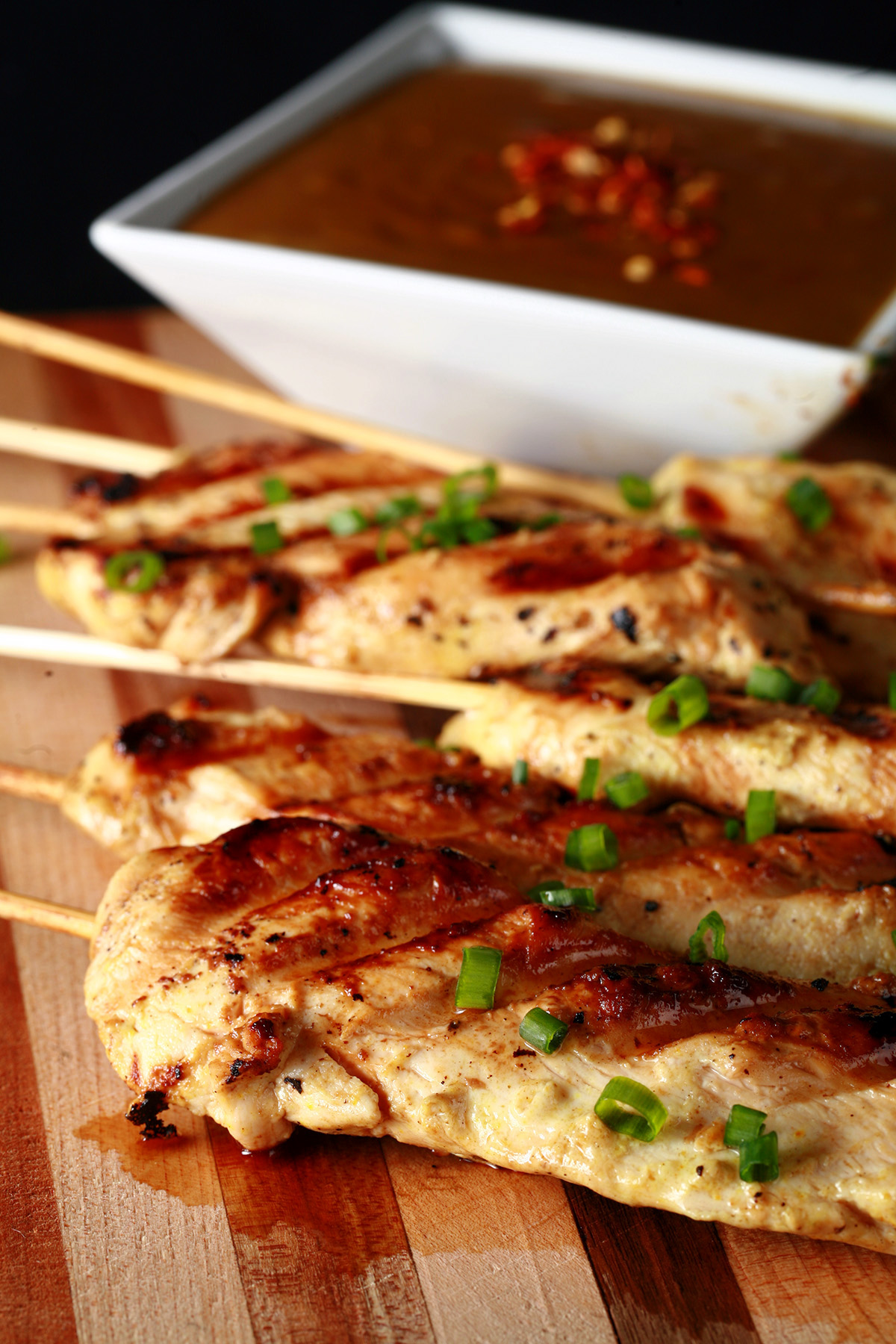 Several skewers of gluten-free chicken satay on a striped cutting board. They are sprinkled with green onions and sitting next to a bowl of peanut sauce.