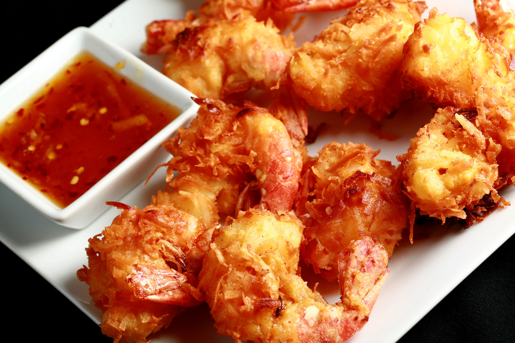 "A white plate is covered with golden brown gluten-free coconut shrimp. There is a small bowl of spicy ginger-orange sauce on the same plate, as a dip.