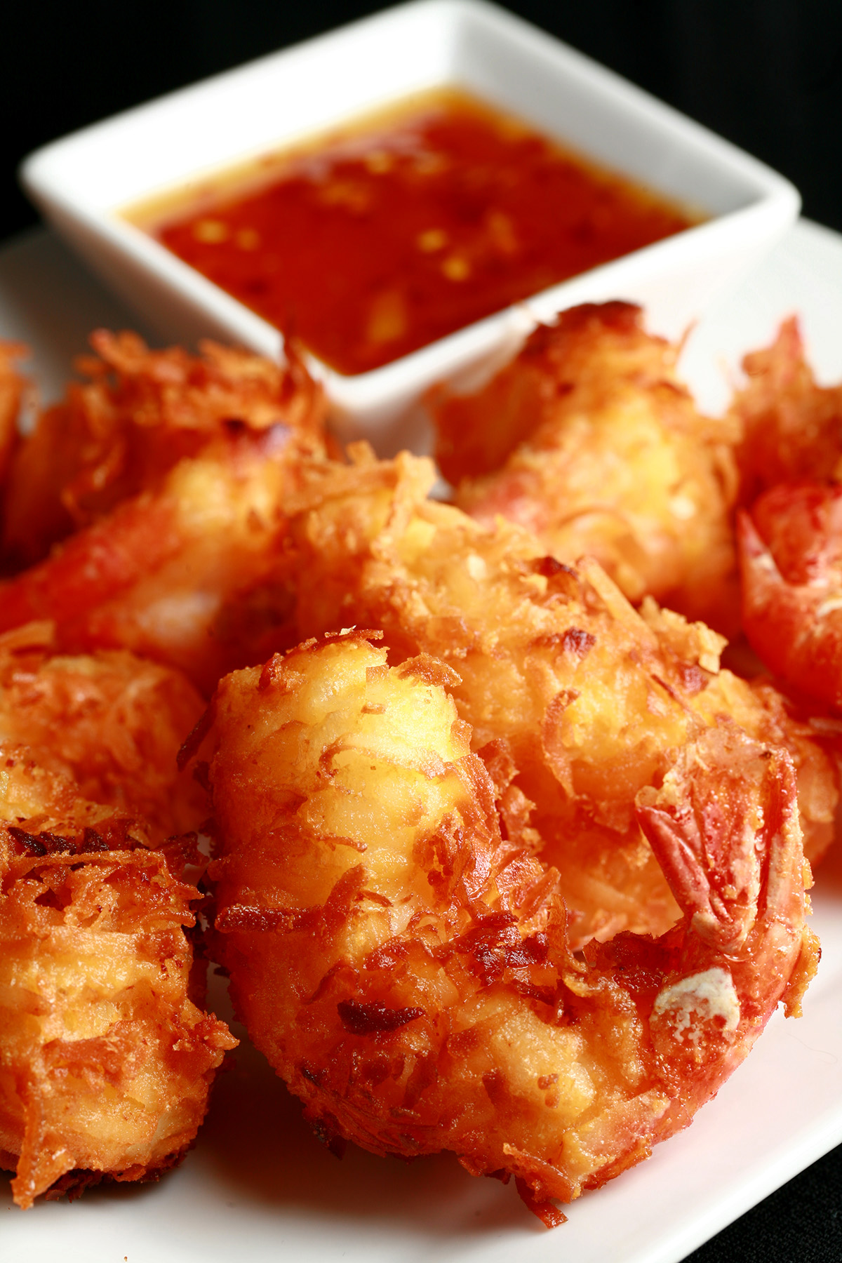 A white plate is covered with golden brown gluten-free coconut shrimp. There is a small bowl of spicy ginger-orange sauce on the same plate, as a dip.