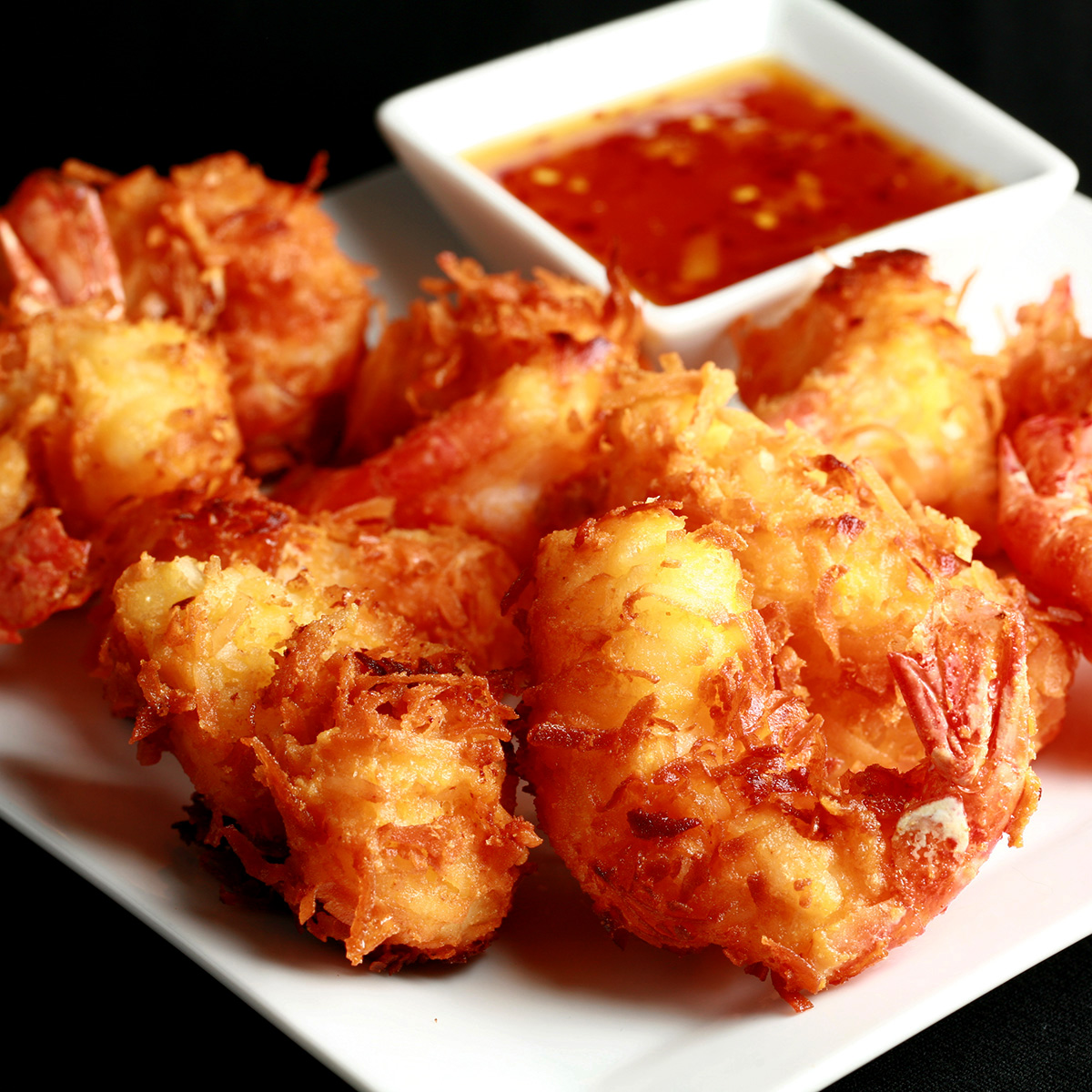 A white plate is covered with golden brown gluten-free coconut shrimp. There is a small bowl of spicy ginger-orange sauce on the same plate, as a dip.