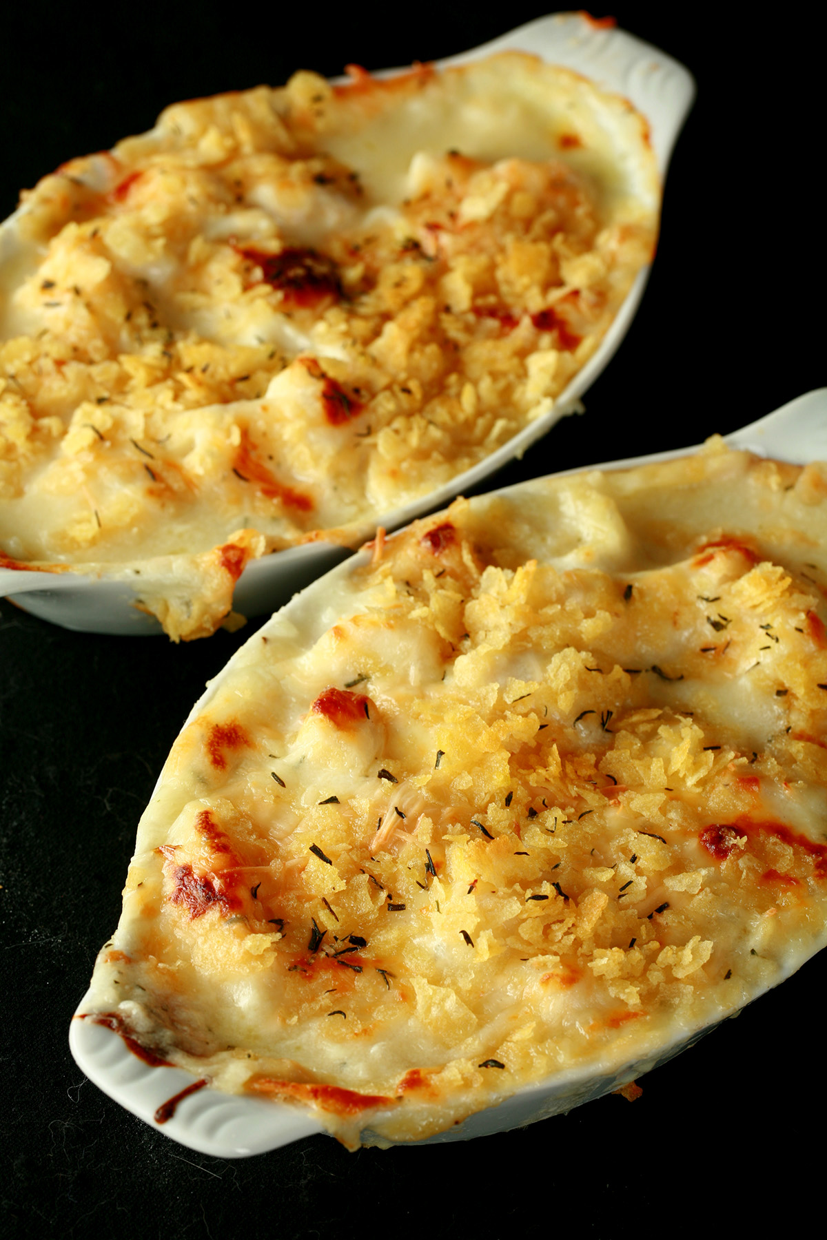 Two oblong ceramic ramekins of gluten-free cod au gratin. Cod is in a creamy, cheesy sauce, topped with crushed chips, more cheese, and savoury.