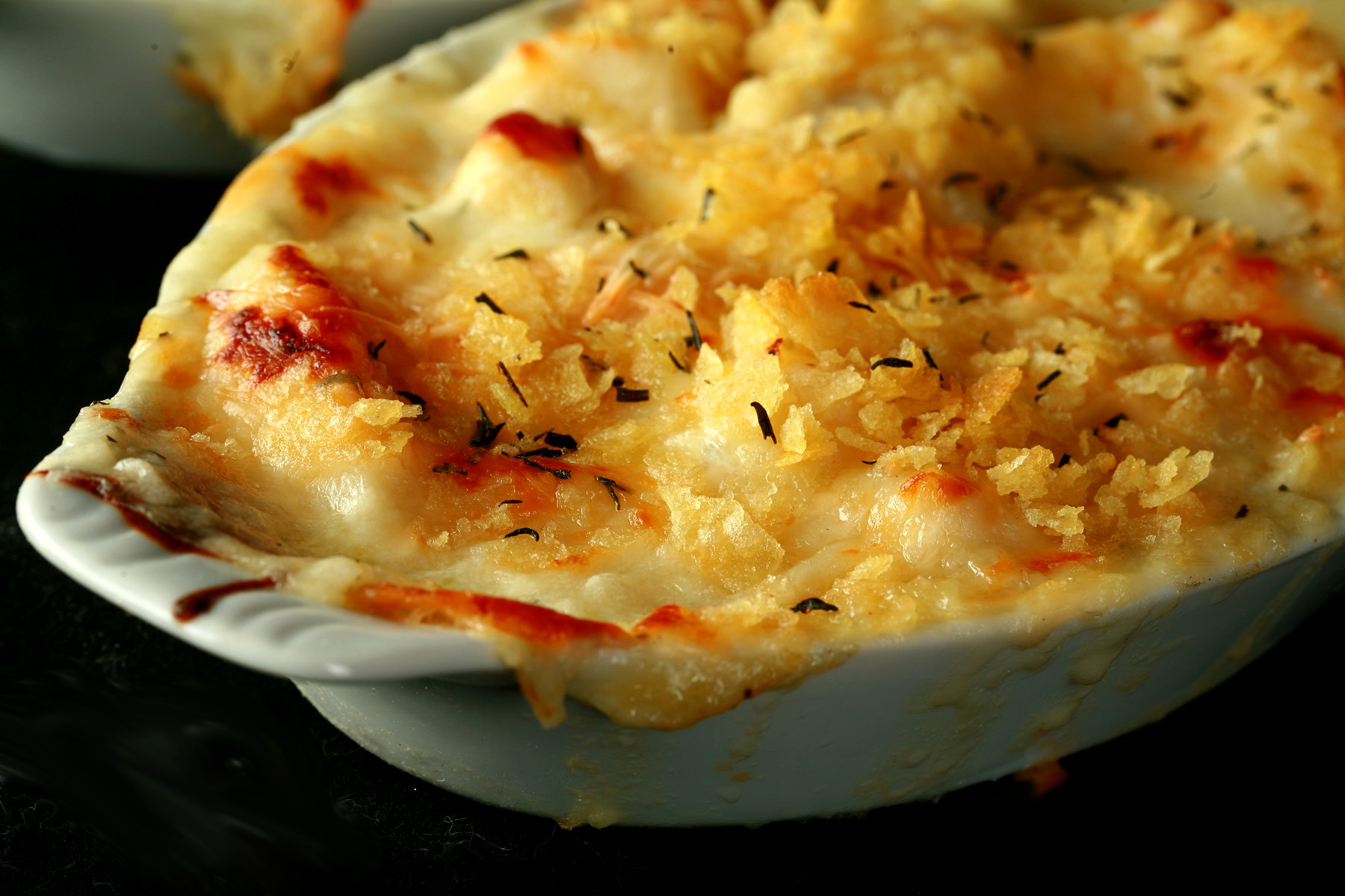 Close up view of a ceramic ramekin of gluten-free cod au gratin. Cod is in a creamy, cheesy sauce, topped with crushed chips, more cheese, and savoury.