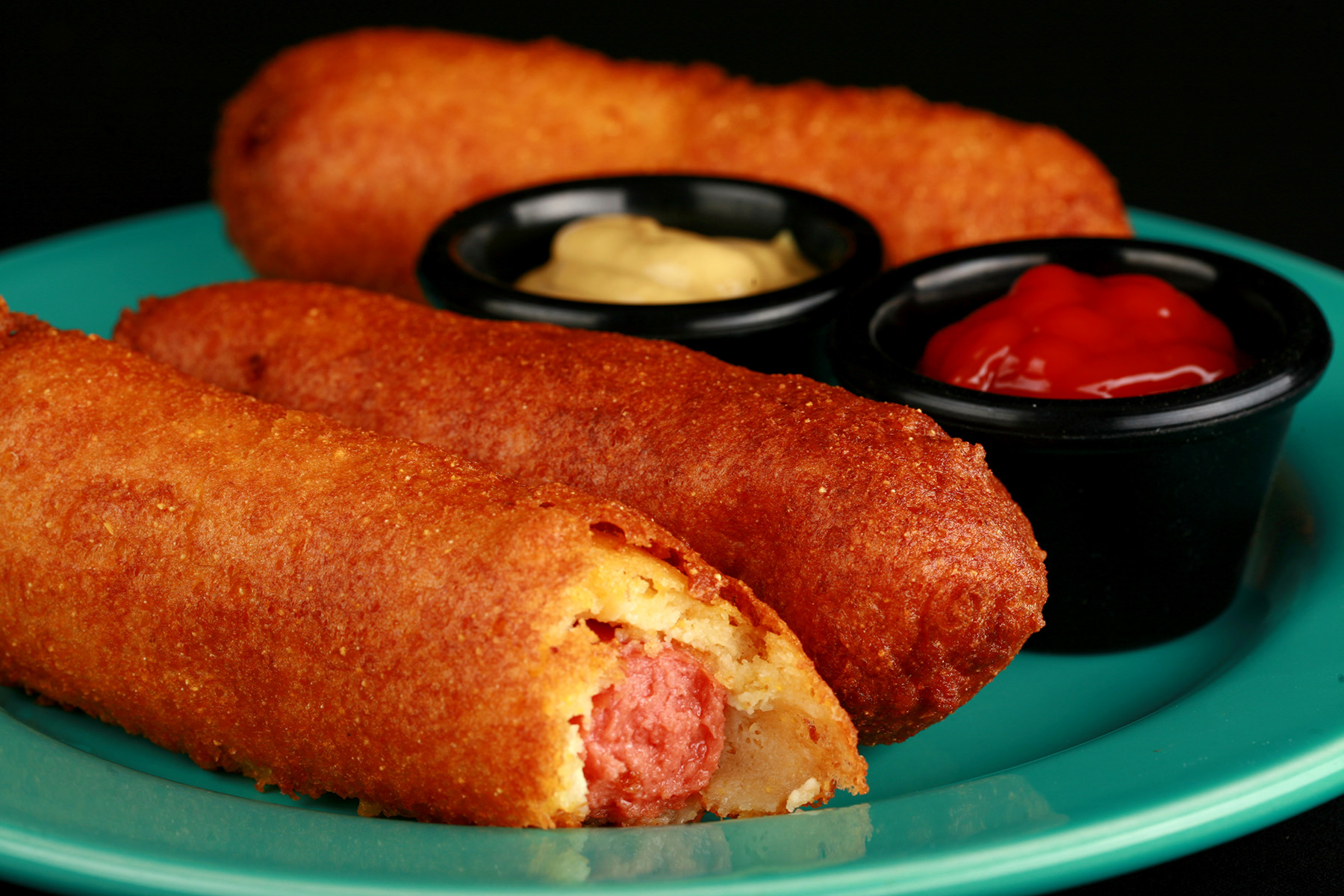 3 gluten-free corndogs on a green plate, with little bowls of ketchup and mustard.