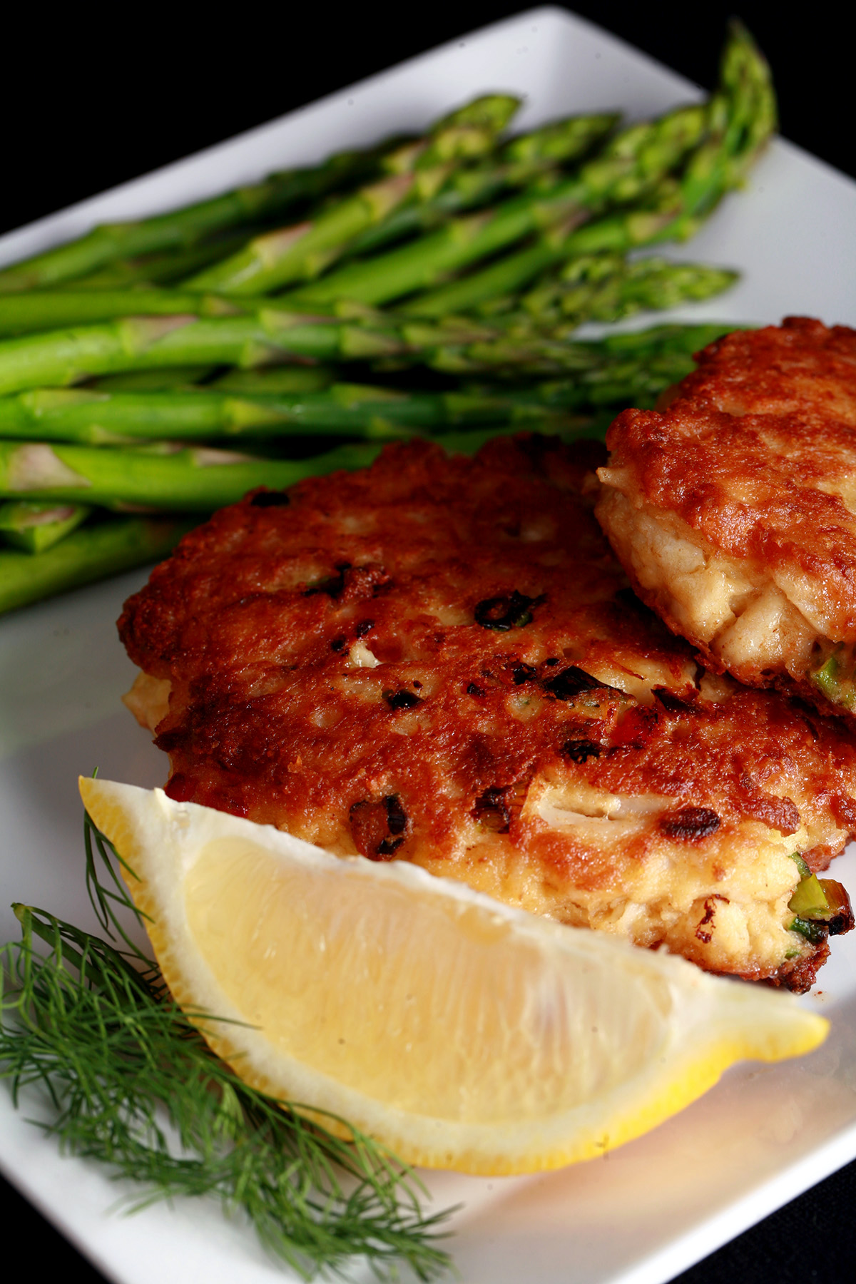 2 Gluten-free Crab Cakes on a plate with asparagus, a lemon slice, and some fresh dill.