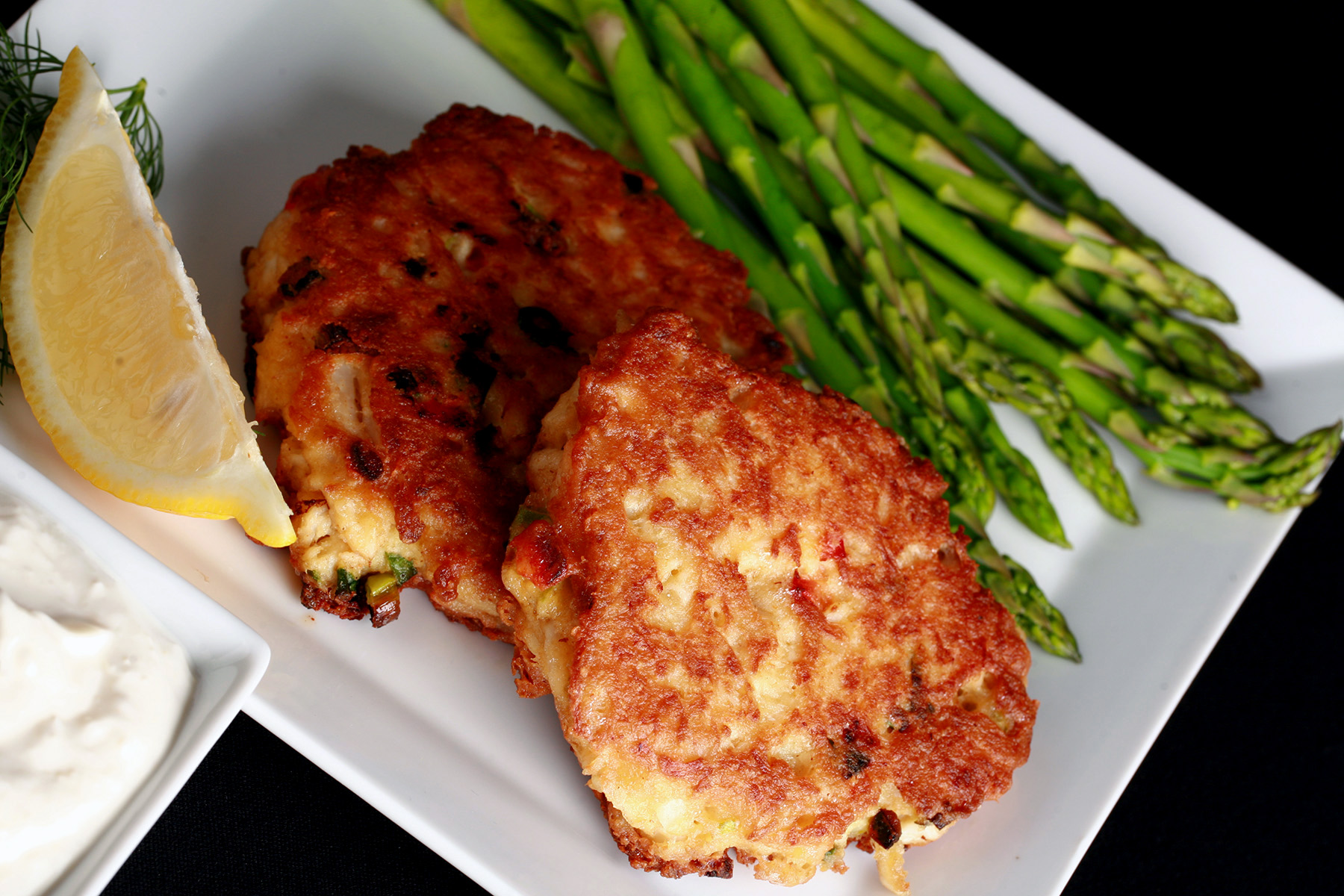2 Gluten-free Crab Cakes on a plate with asparagus, a lemon slice, and some fresh dill. There is a small bowl of horseradish sauce next to it.