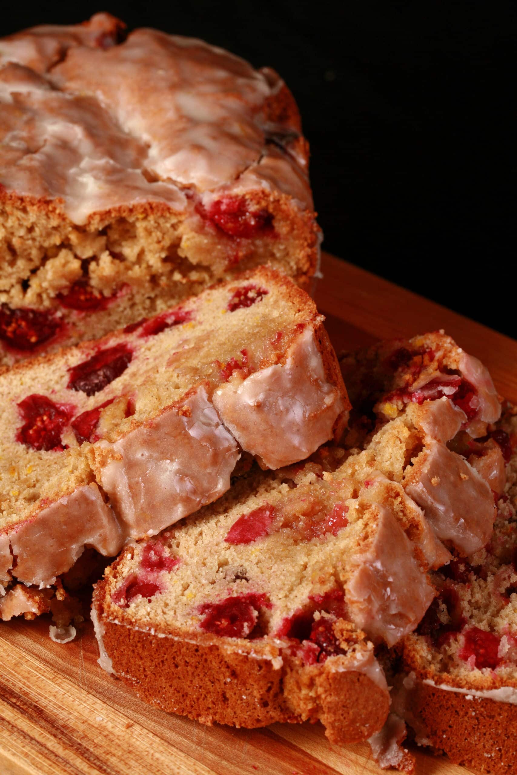 A loaf of glazed gluten free orange cranberry bread, sliced on a cutting board.