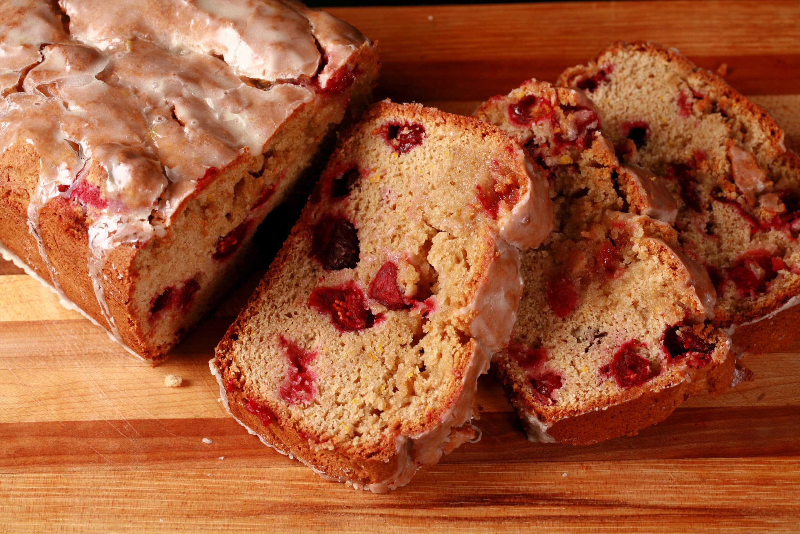 A loaf of glazed gluten free orange cranberry bread, sliced on a cutting board.