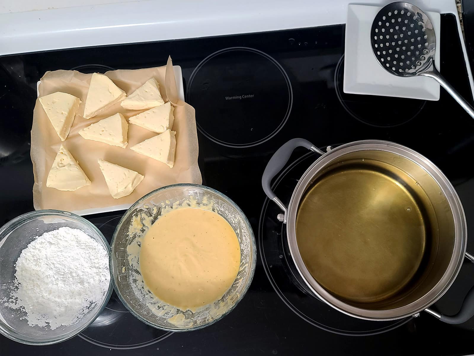 A plate of brie wedges, a bowl of corn starch, a bowl of batter, and a pot of oil on a stove top.