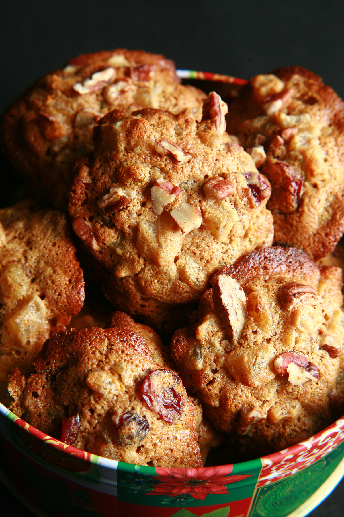 A festive green and red holiday container is full of cookies. The cookies have visible pieces of fruits and nuts throughout.