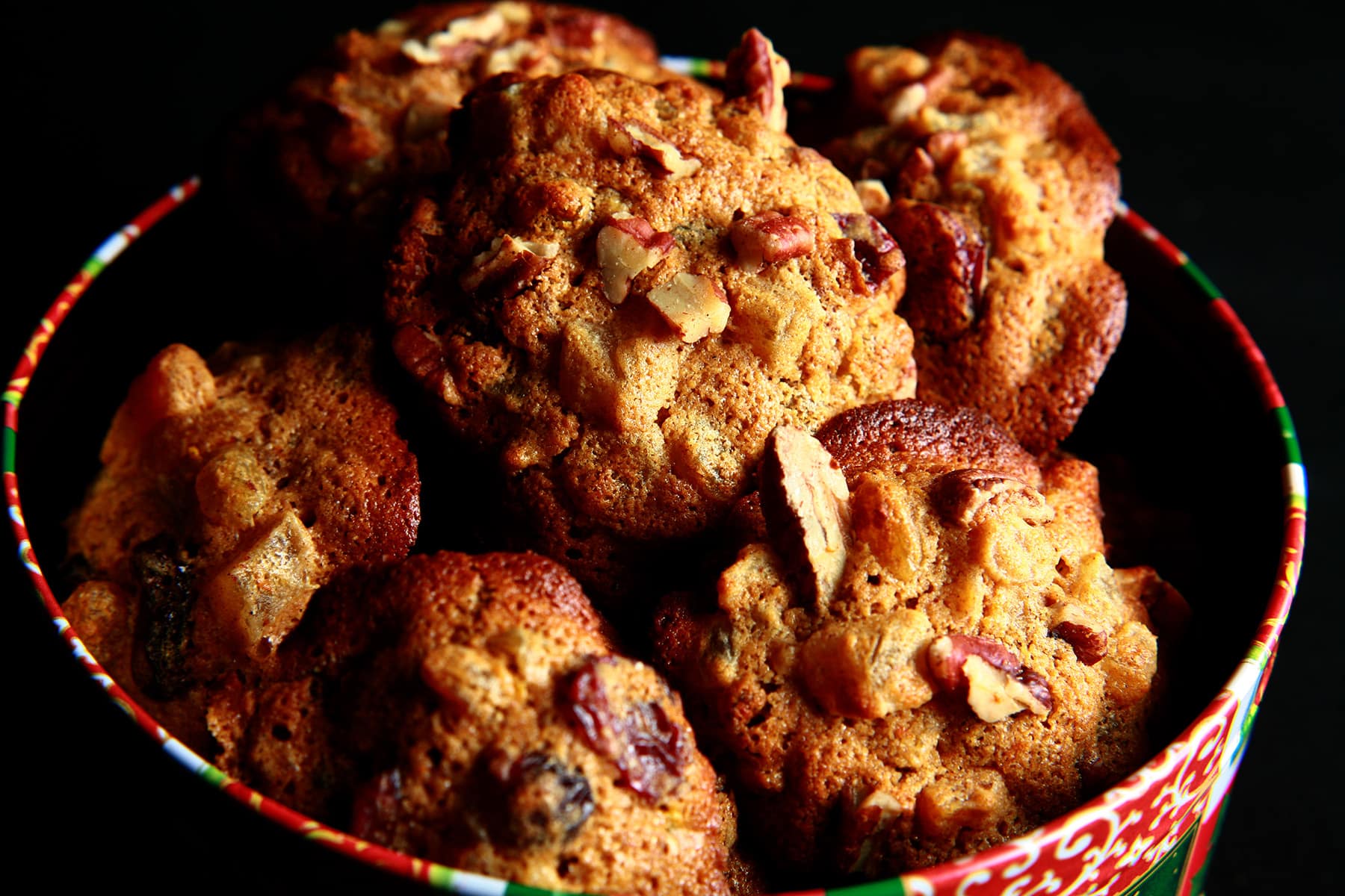 A festive green and red holiday container is full of cookies. The cookies have visible pieces of fruits and nuts throughout.