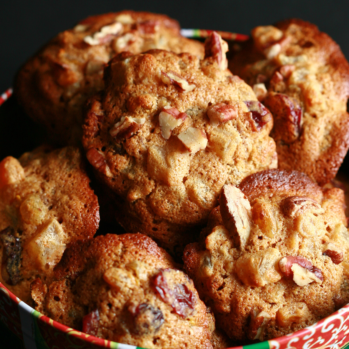 A festive green and red holiday container is full of cookies. The cookies have visible pieces of fruits and nuts throughout.