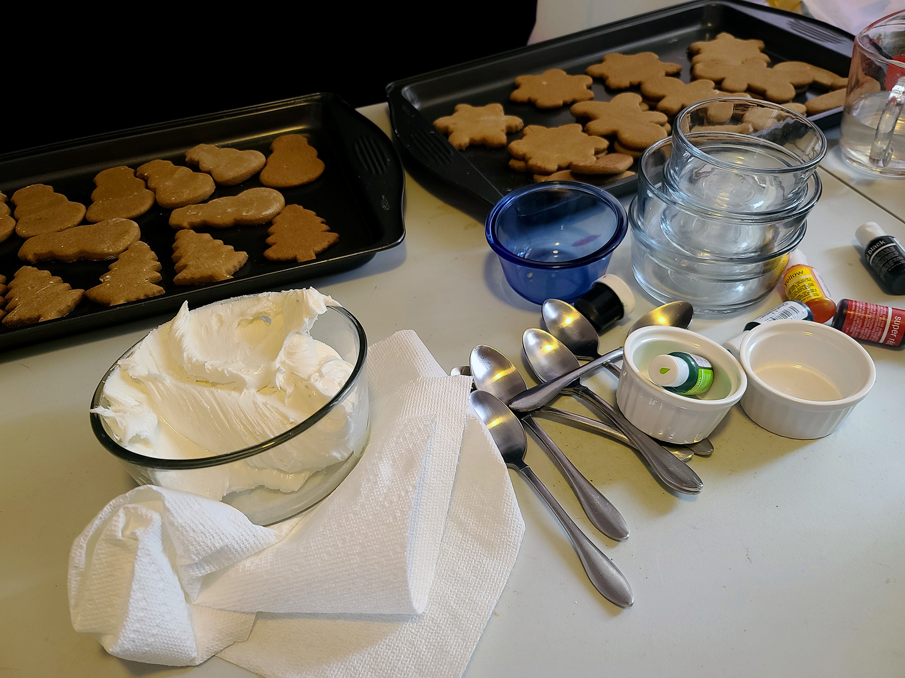 A work surface with cookies, a bowl of frosting, food colouring, and spoons.