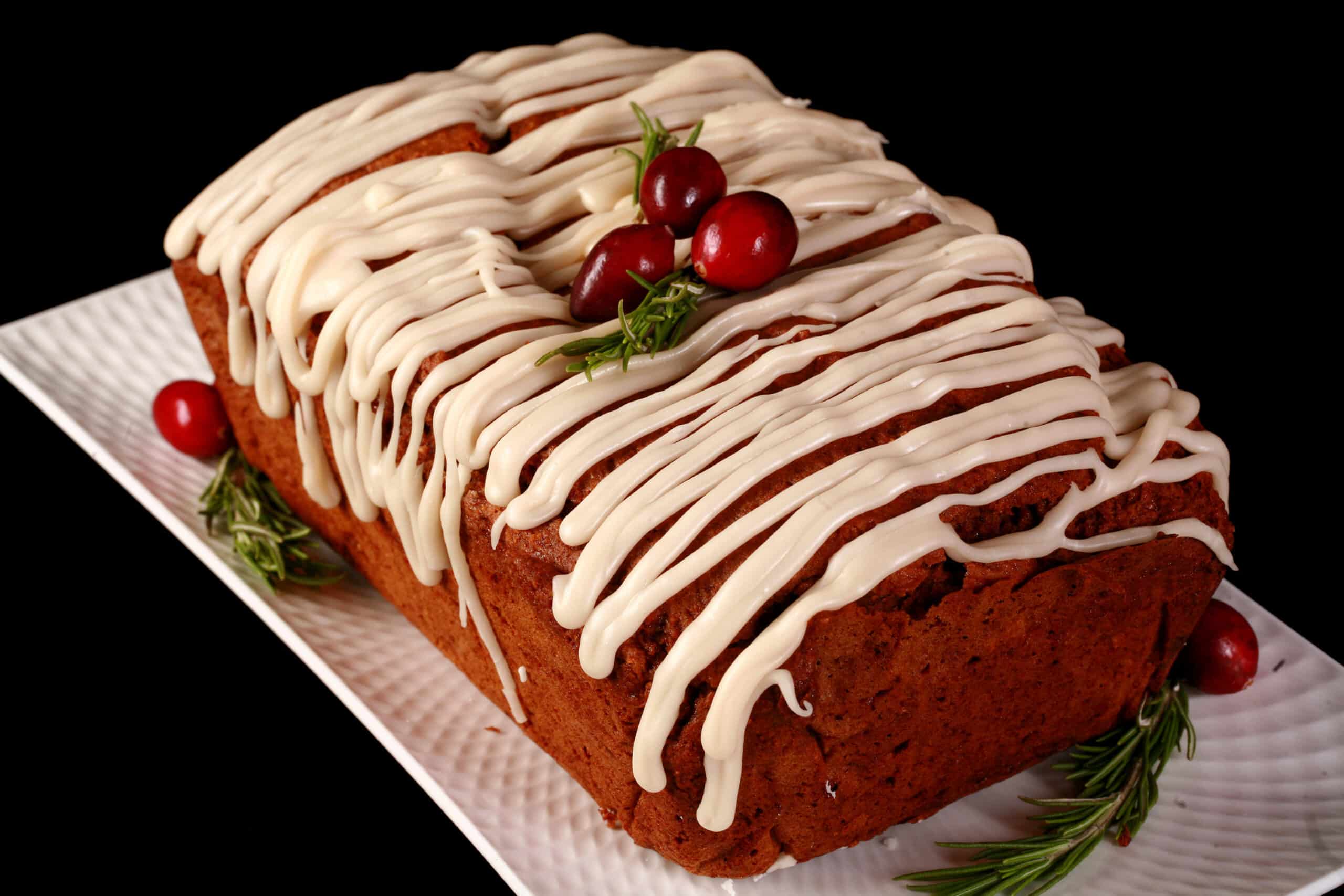 A loaf of gluten free gingerbread loaf, drizzled with vanilla glaze and garnished with rosemary and cranberries.