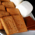 A white plate with rows of square shaped gluten-free graham crackers, a couple large marshmallows, and a piece of chocolate.