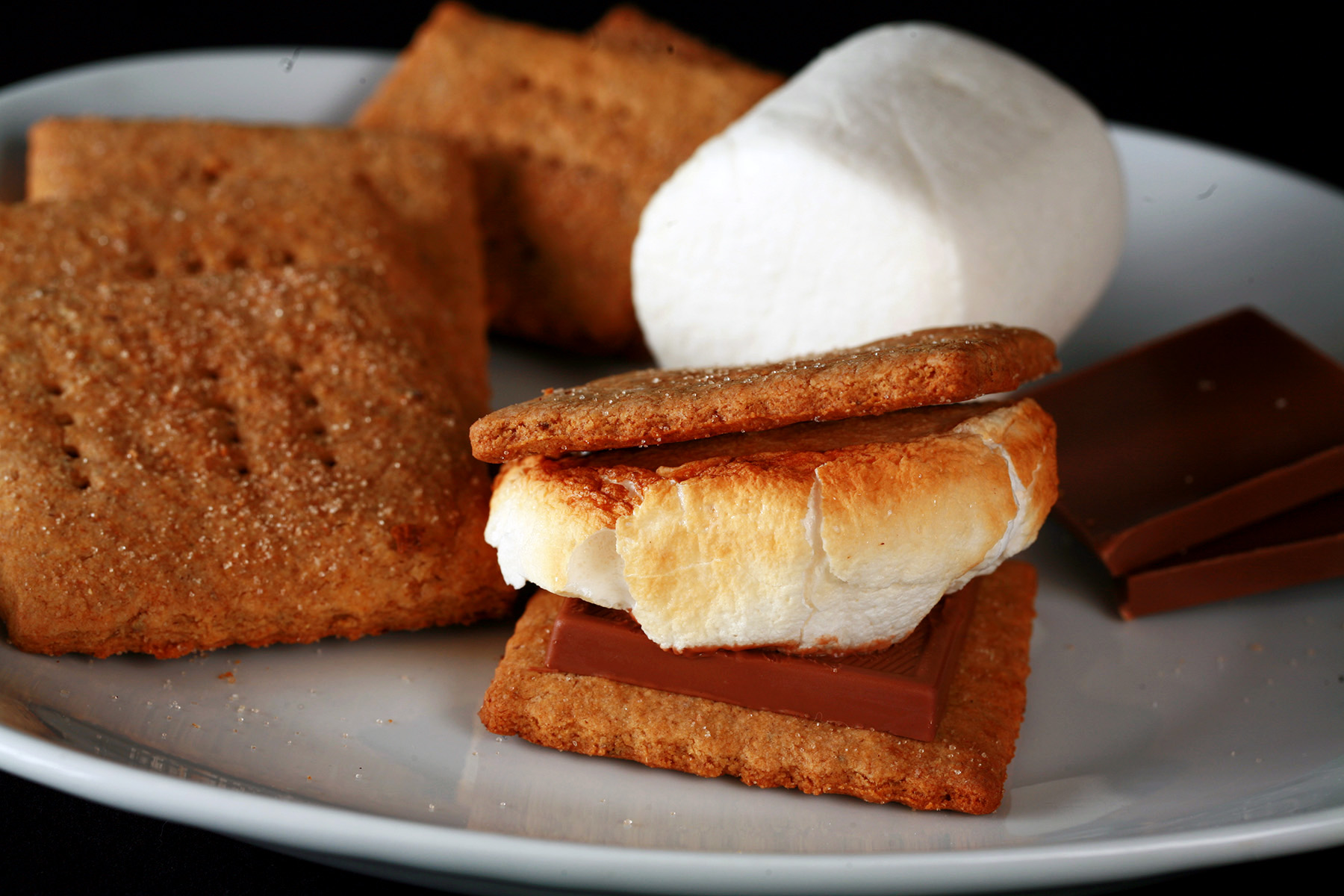 A white plate with rows of square shaped gluten-free graham crackers, a couple large marshmallows, and a piece of chocolate. Two crackers,a piece of chocolate, and a toasted marshmallow have been assembled into a S'more also on the place.