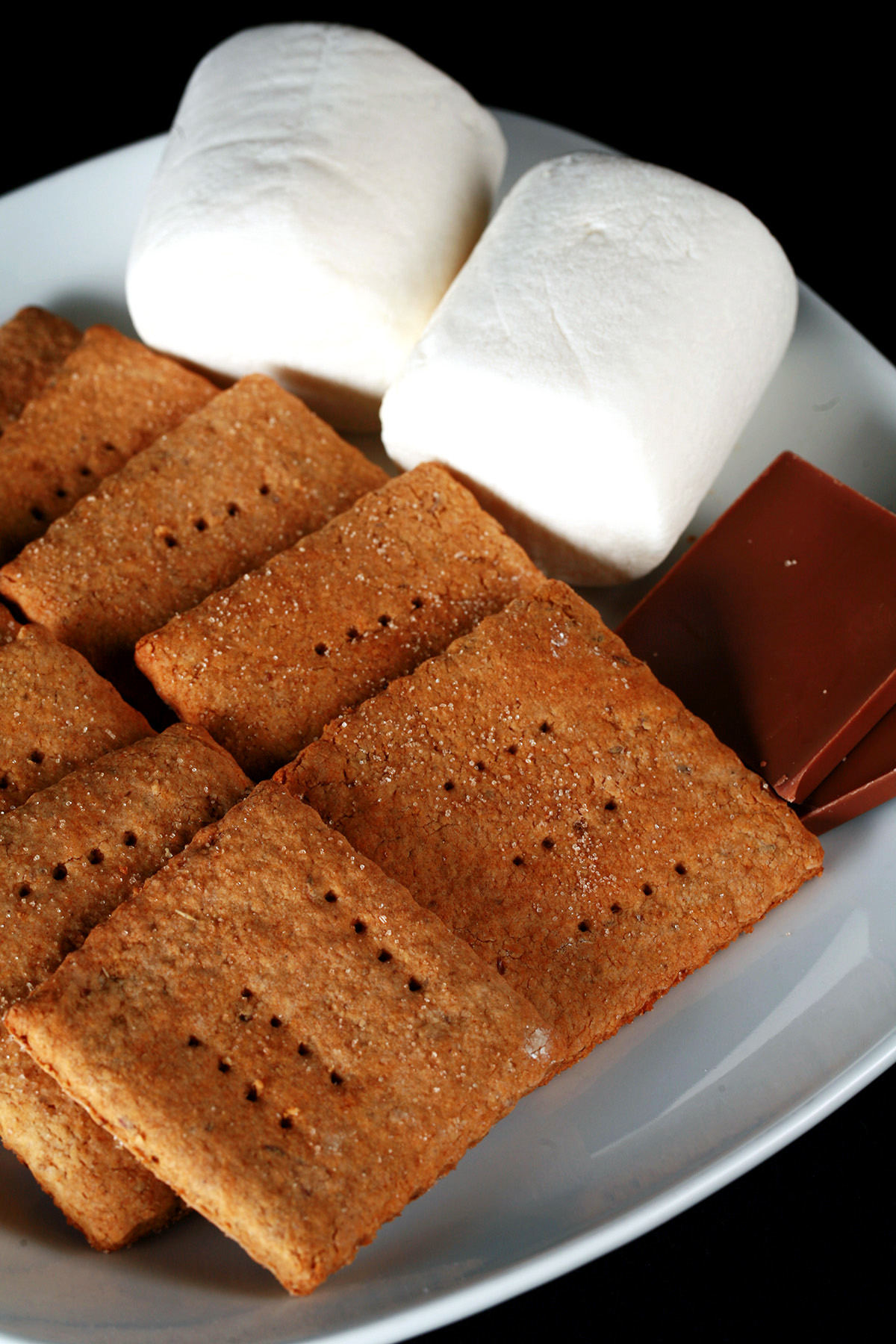 A white plate with rows of square shaped gluten-free graham crackers, a couple large marshmallows, and a piece of chocolate.