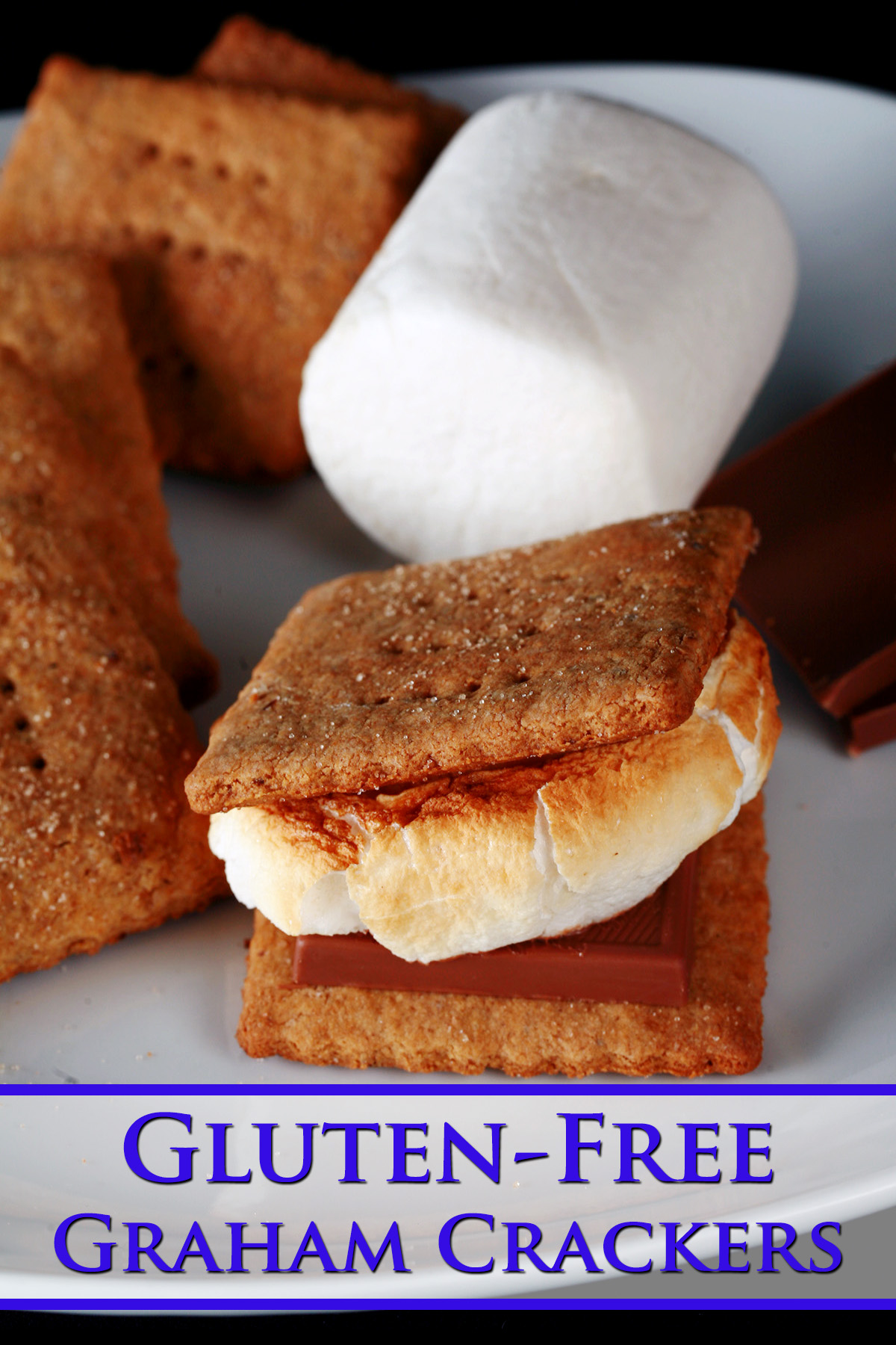 A white plate with rows of square shaped gluten-free graham crackers, a couple large marshmallows, and a piece of chocolate. Two crackers,a  piece of chocolate, and a toasted marshmallow have been assembled into a s’more also on the plate.