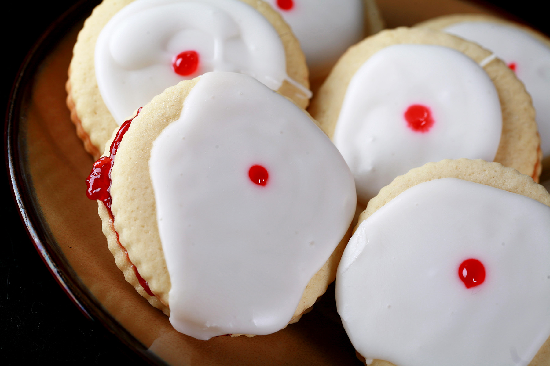 Close up view of a plate of Gluten-Free Empire Cookies - sandwich cookies filled with raspberry jam, frosted with a white glaze, and finished off with a dot of red gel in the center.