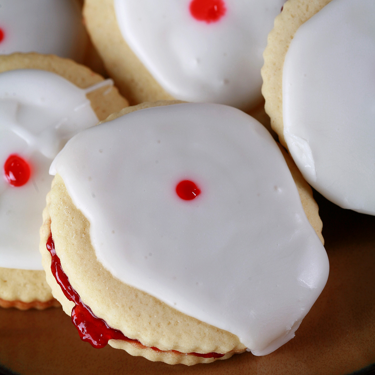 "Close up view of a plate of Gluten-Free Empire Cookies - sandwich cookies filled with raspberry jam, frosted with a white glaze, and finished off with a dot of red gel in the center.