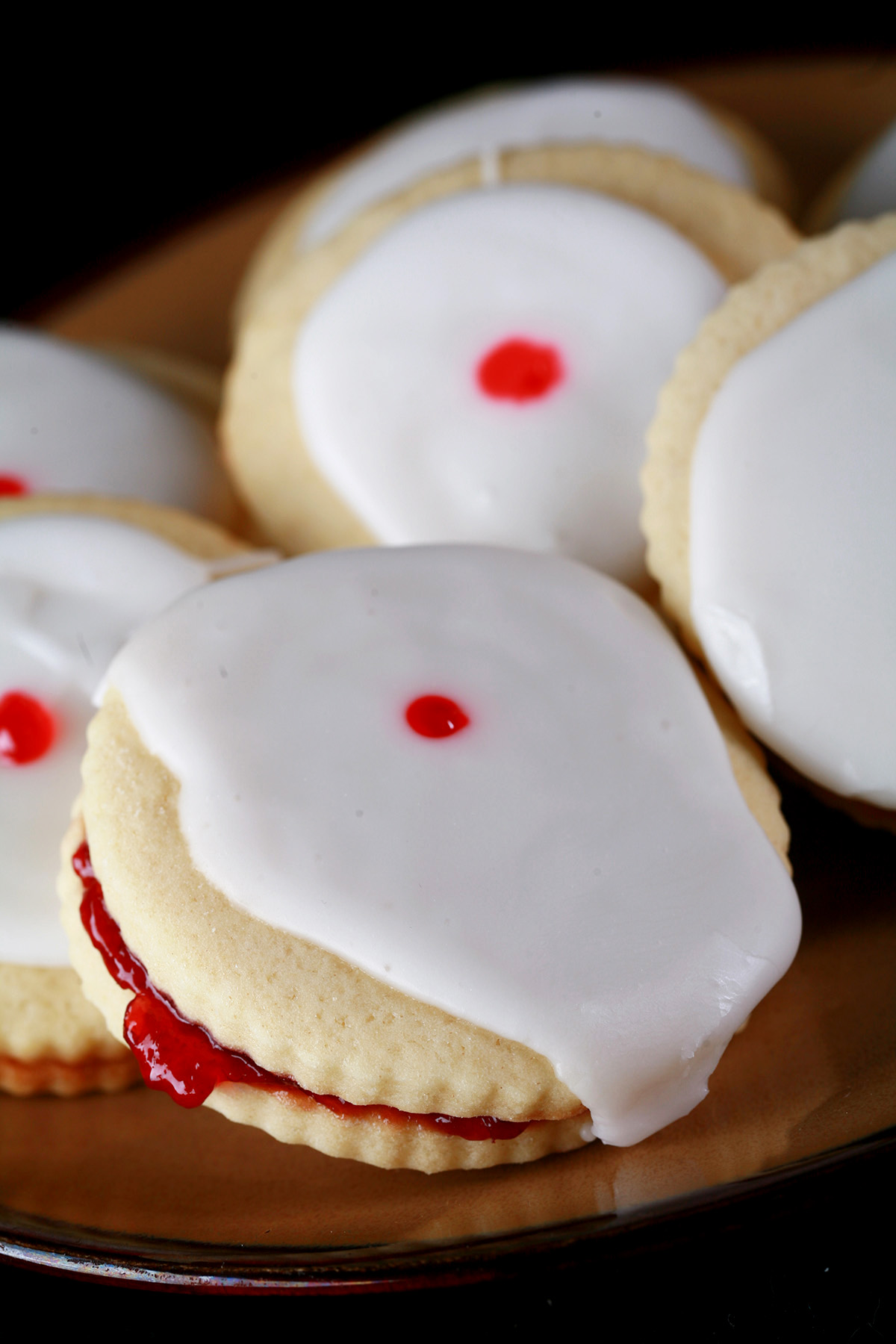 Close up view of a plate of Gluten-Free Empire Biscuits - sandwich cookies filled with raspberry jam, frosted with a white glaze, and finished off with a dot of red gel in the center.