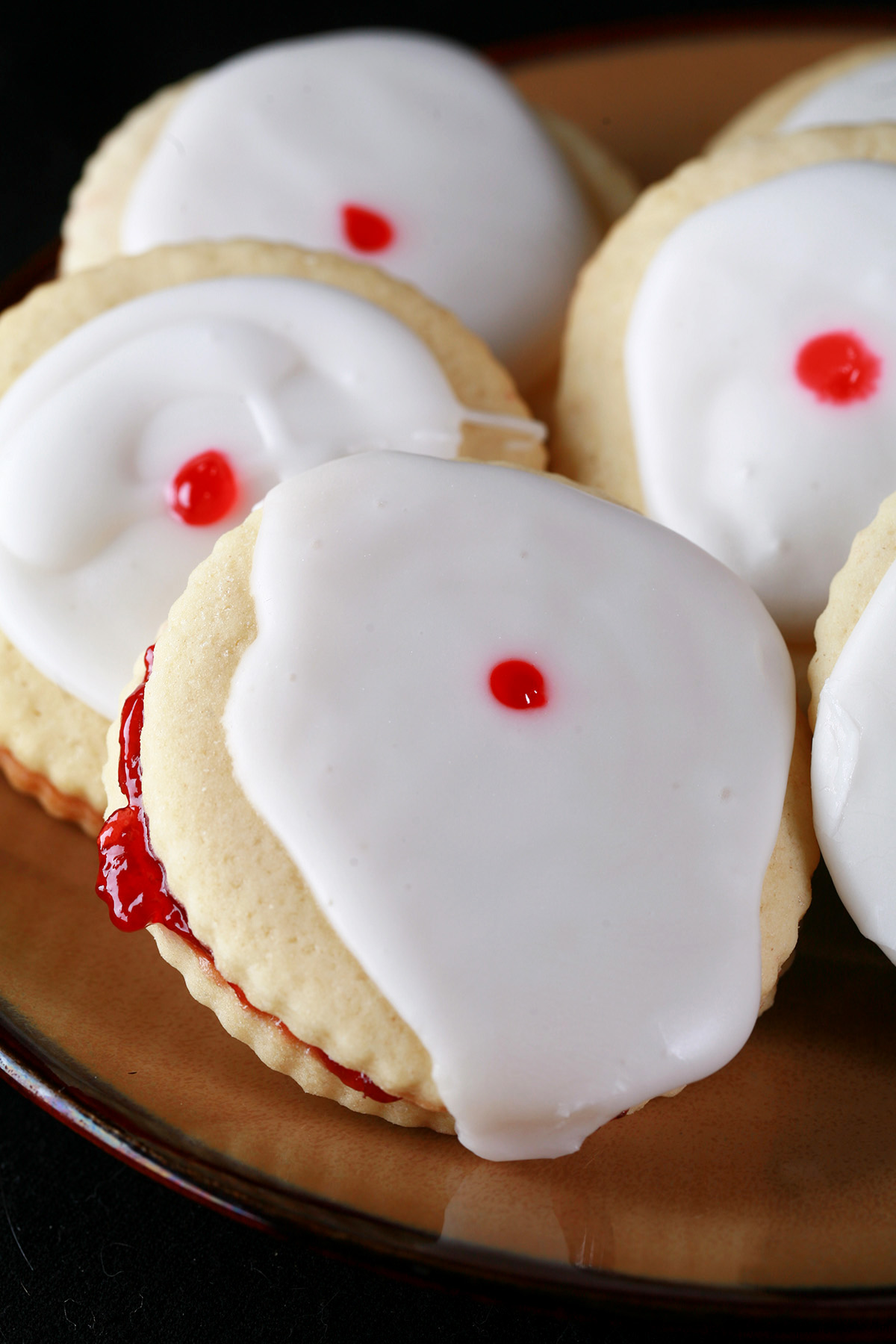 Close up view of a plate of Gluten-Free Empire Cookies - sandwich cookies filled with raspberry jam, frosted with a white glaze, and finished off with a dot of red gel in the center.