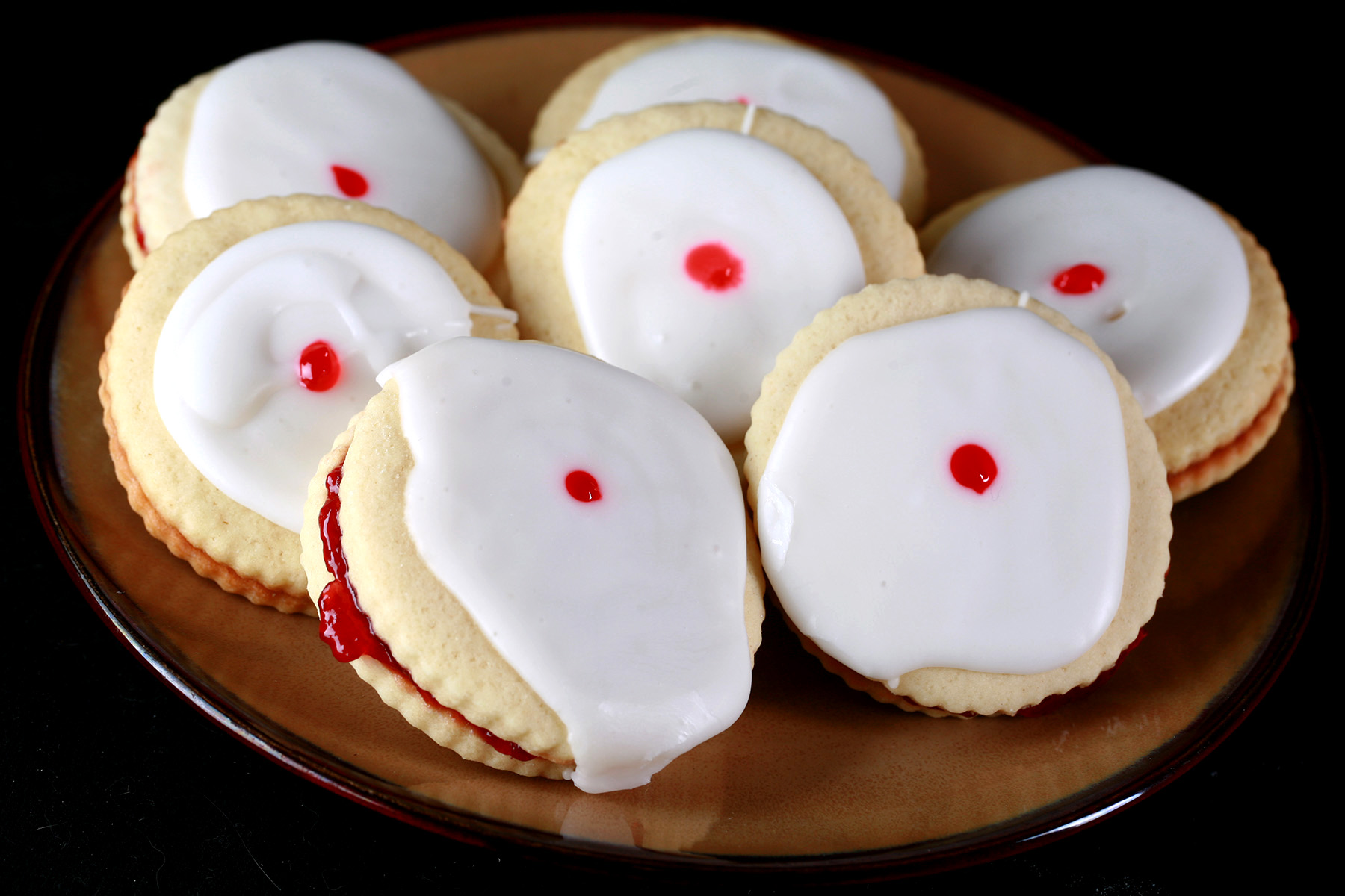 "Close up view of a plate of gluten-free imperial cookies: Sandwich cookies filled with raspberry jam, frosted with a white glaze, and finished off with a dot of red gel in the center.