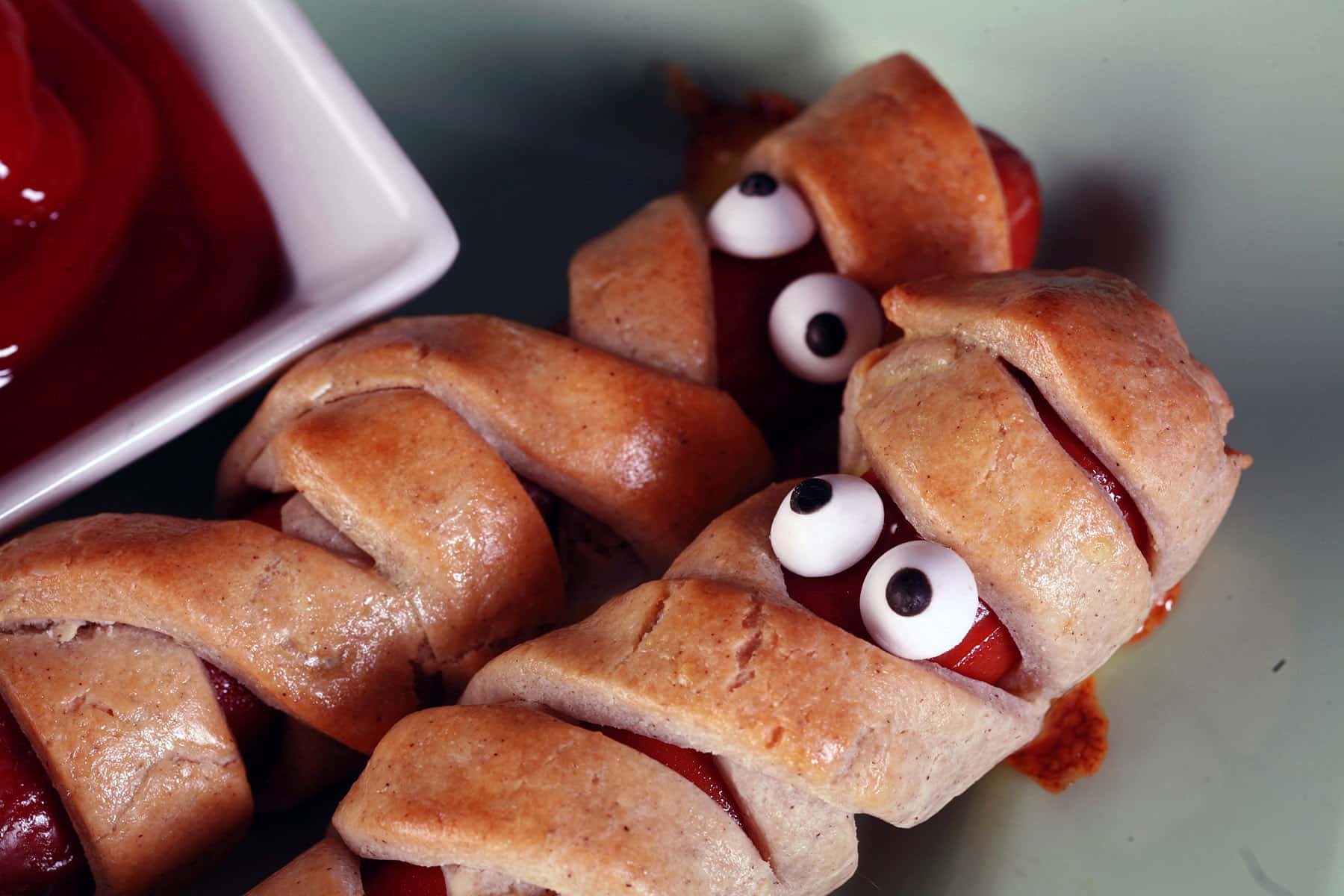 Several gluten-free mummy dogs on a plate. Each have 2 candy eyes.