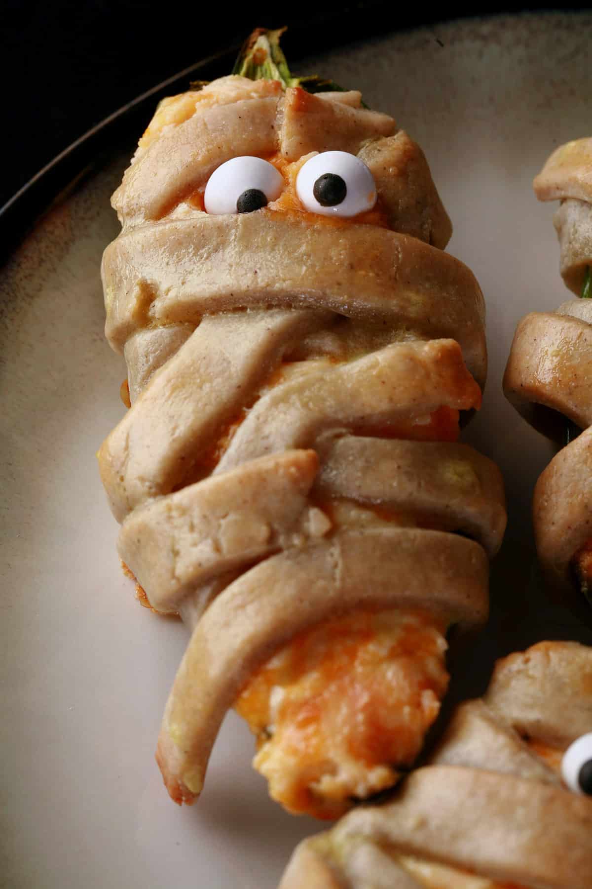 Several gluten free mummy jalapeno poppers on a plate. Each has 2 candy eyeballs.