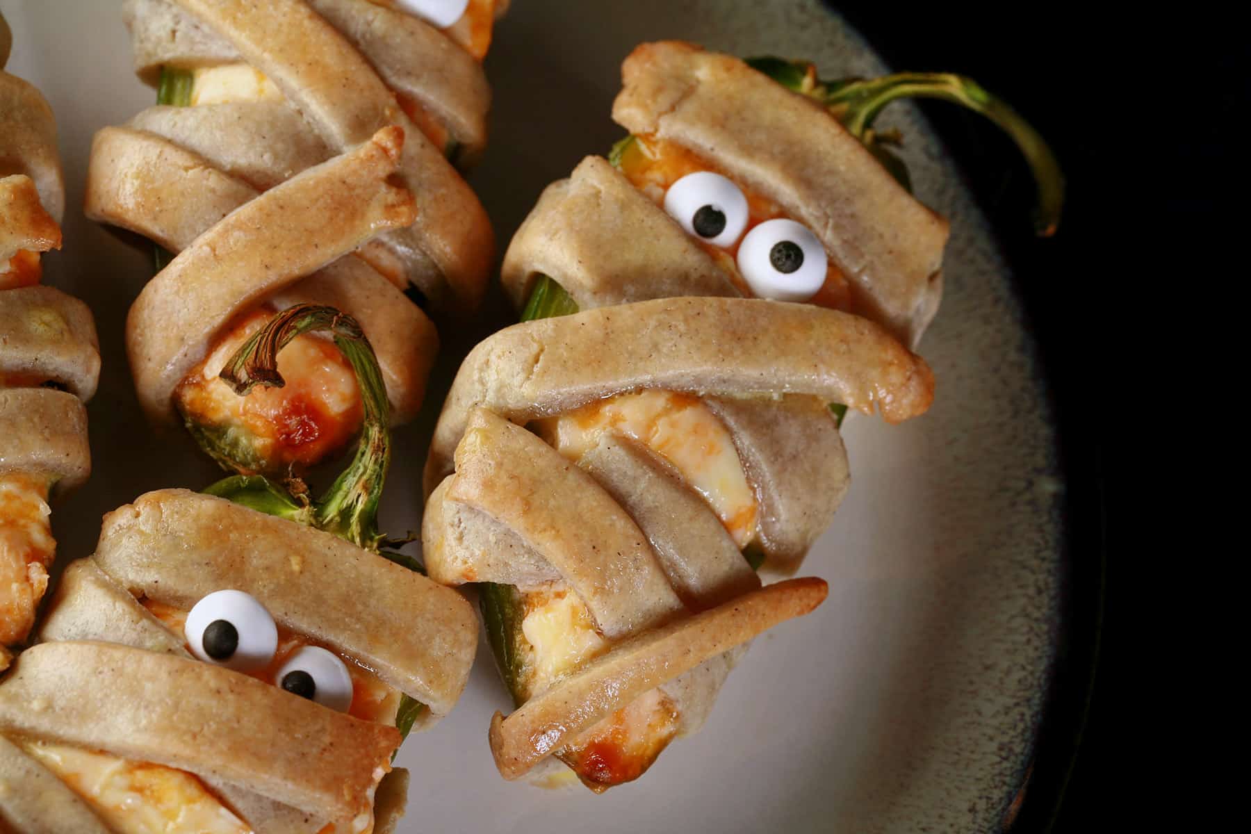 Several gluten free mummy jalapeno poppers on a plate. Each has 2 candy eyeballs.